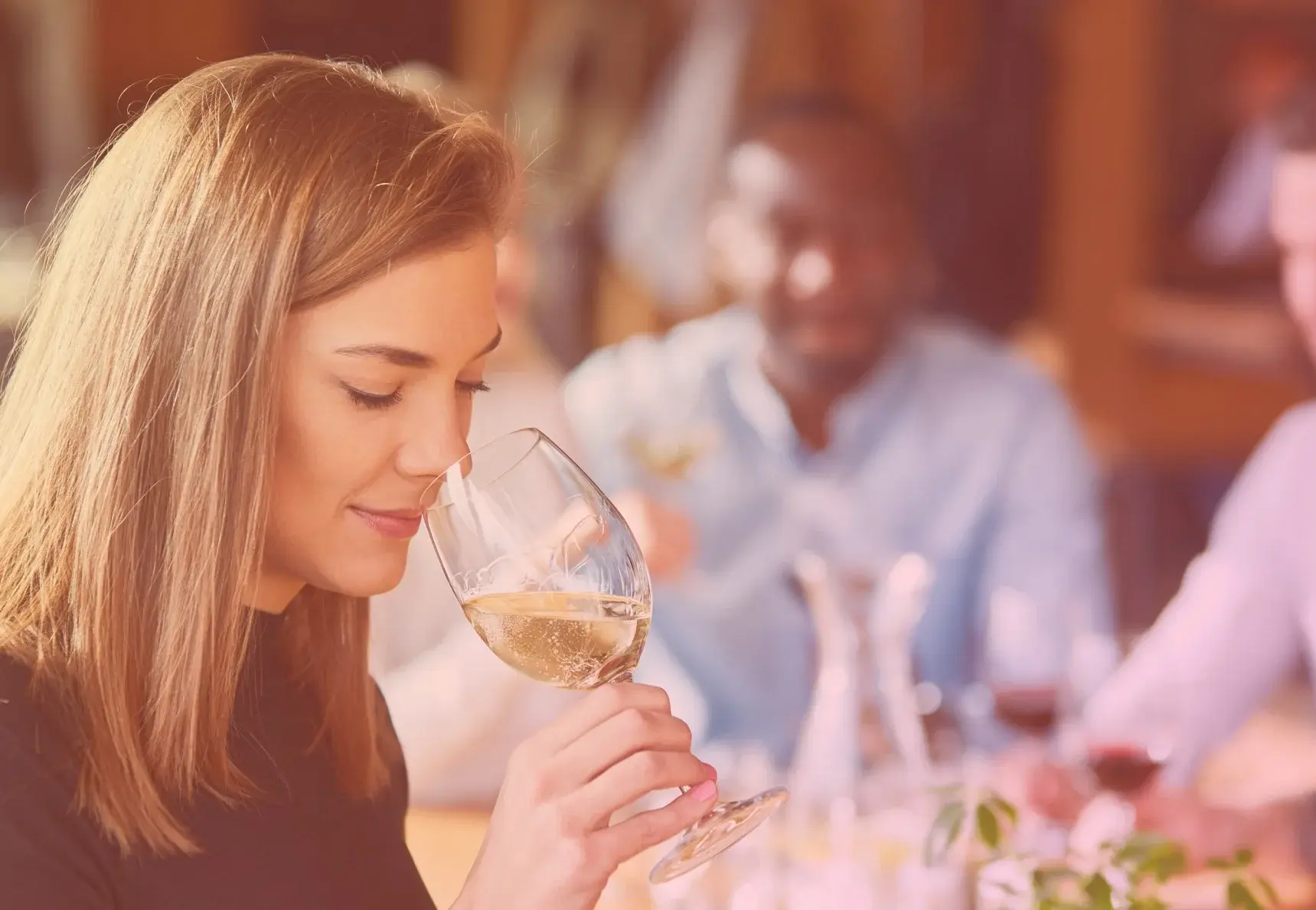 Donna che annusa il vino bianco con persone sullo sfondo.
