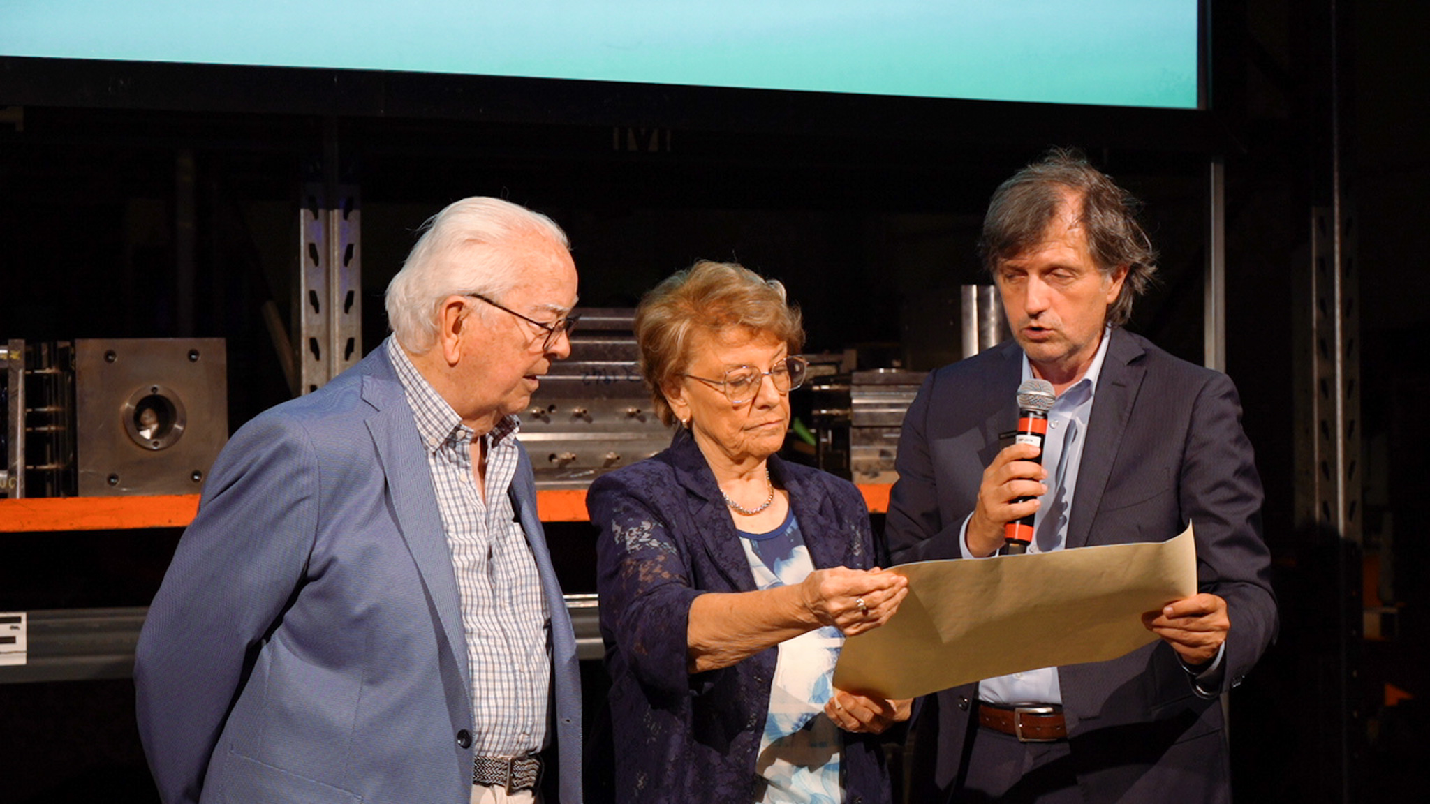 Tre persone in piedi su un palco, una donna sta esaminando un documento.
