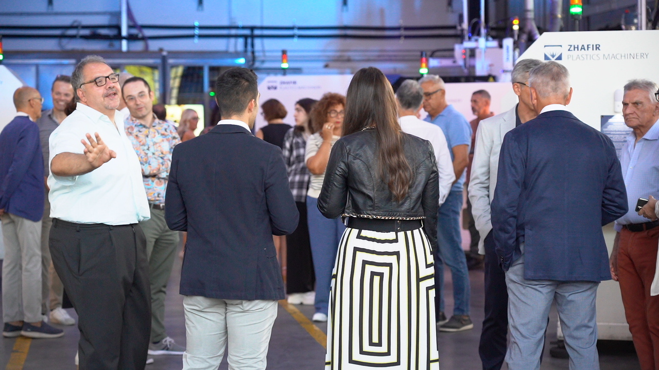 Persone in piedi in un edificio con la scritta "Zhafir Plastics Machinery".