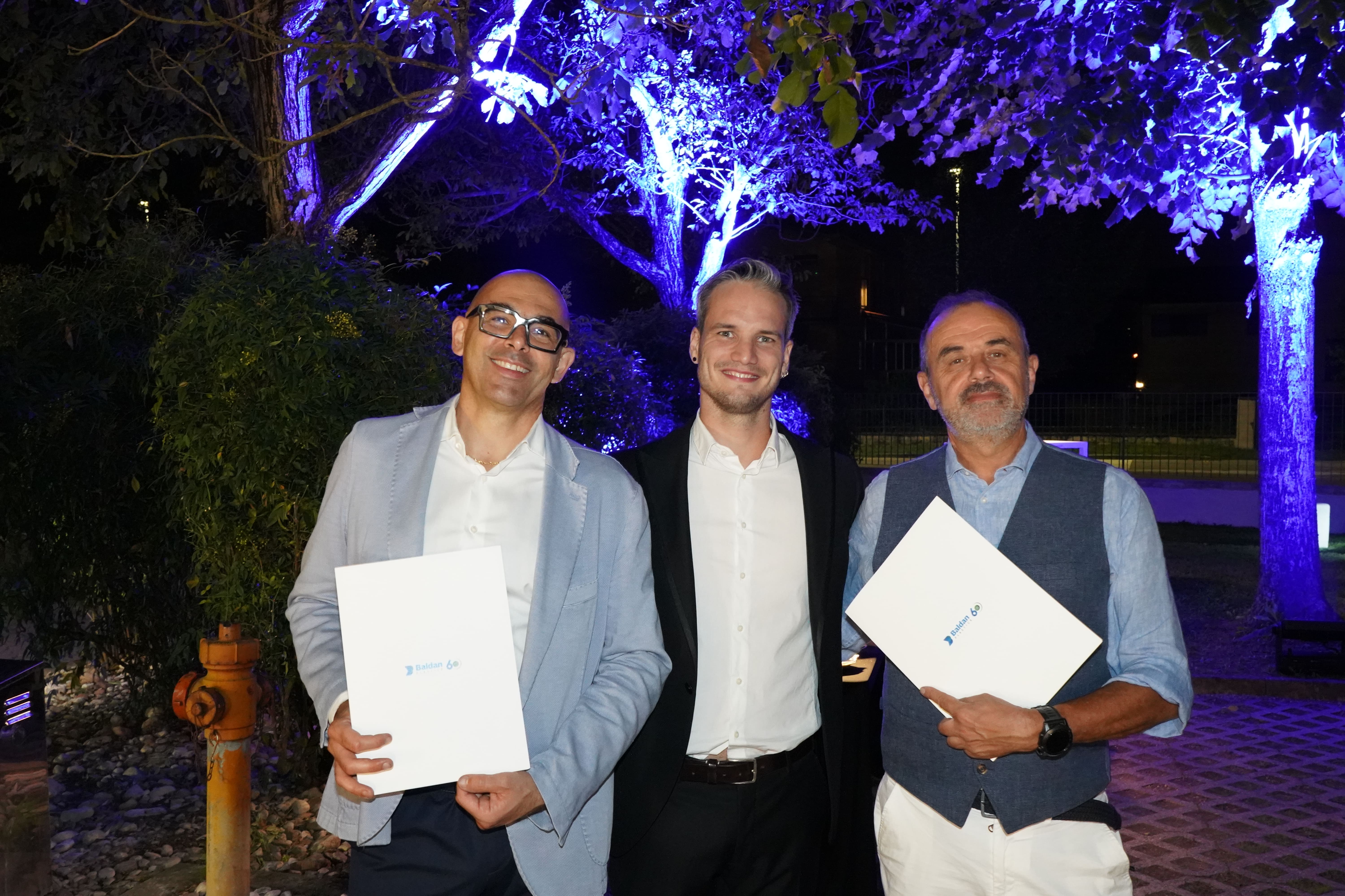 Tre uomini che sorridono e tengono in mano dei documenti di "Baldan 60".