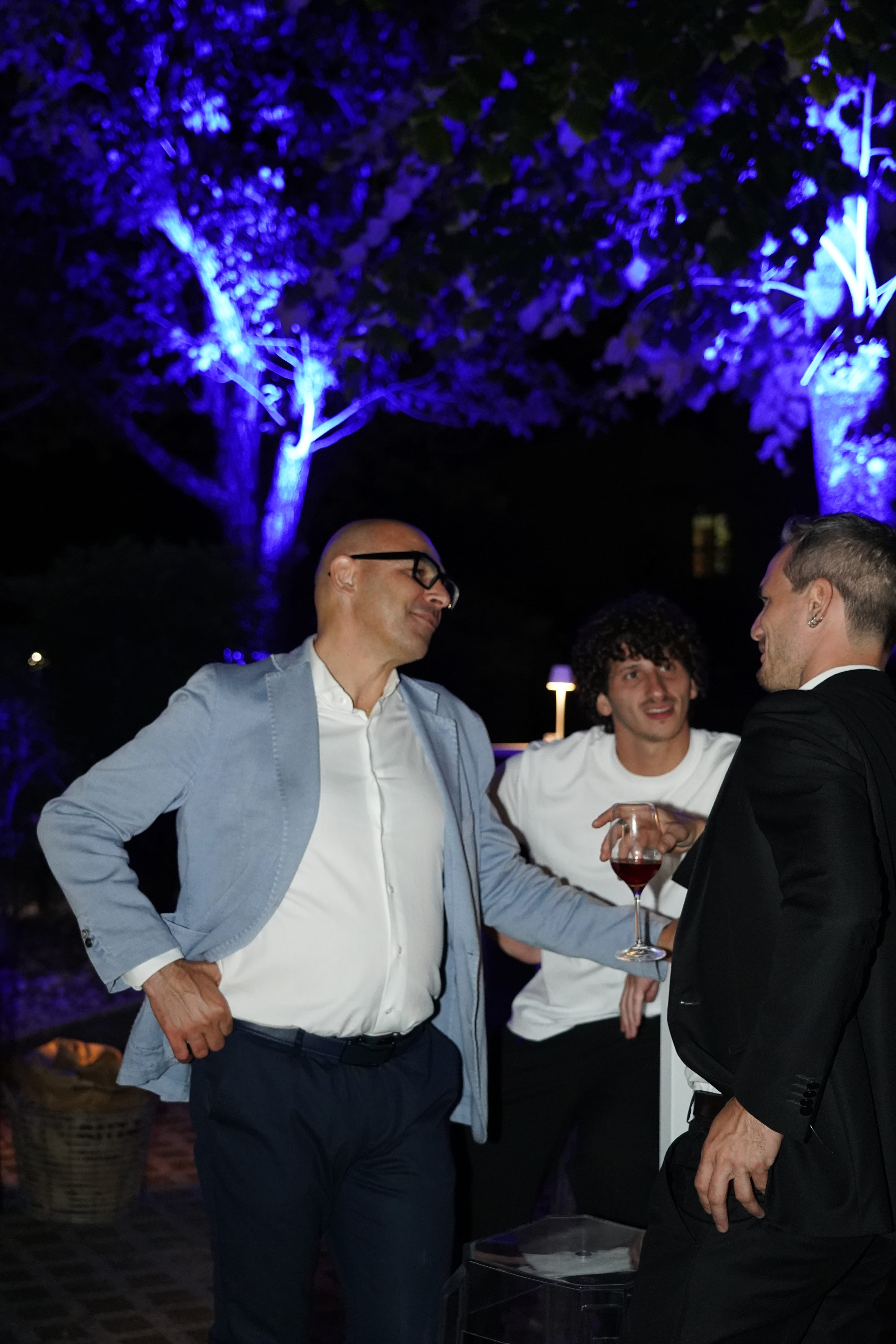 Tre uomini che chiacchierano a una festa all'aperto sotto un albero illuminato di blu.