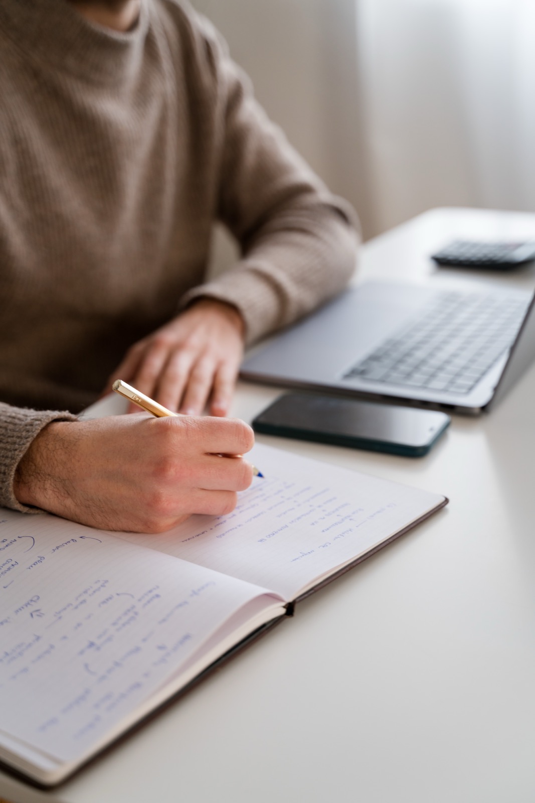 A hand writing in a notebook next to a laptop on a desk