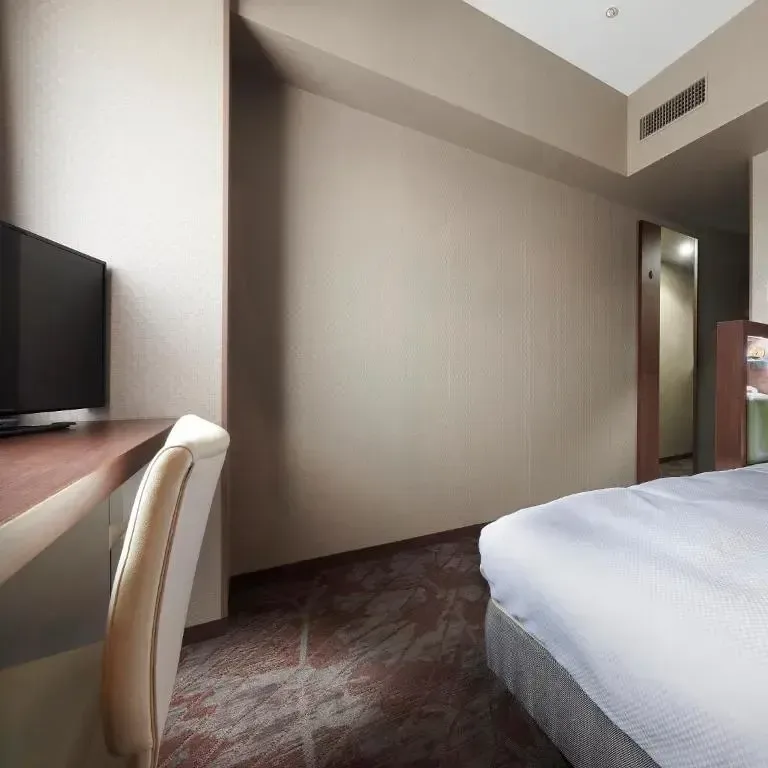 Clean and modern hotel bedroom featuring a made bed, desk with a television, and neutral walls.