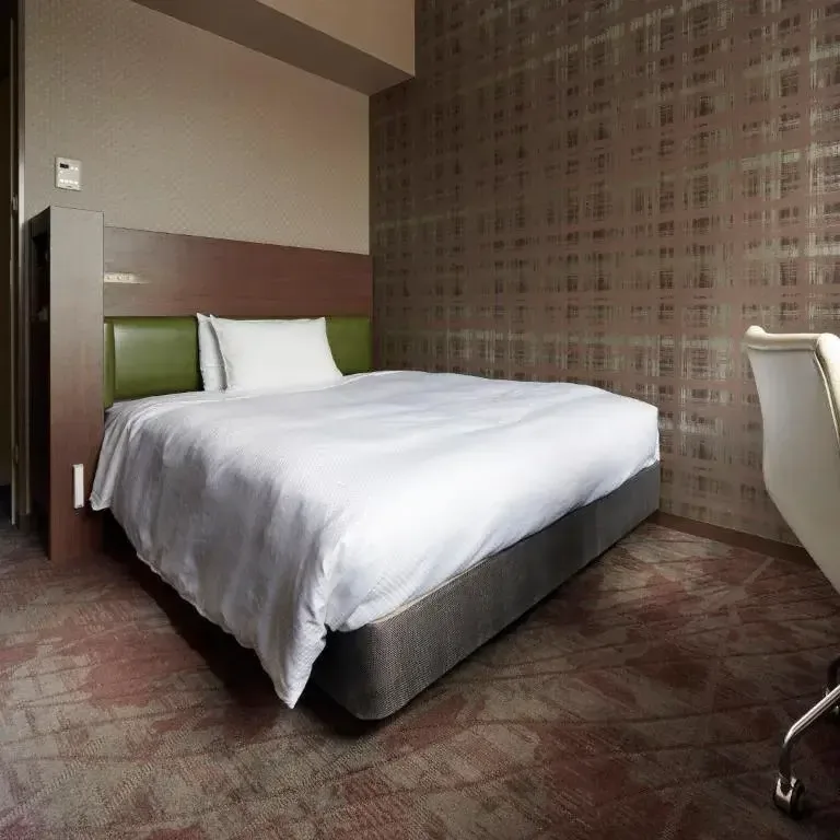 Modern hotel room interior featuring a made double bed with white bedding, patterned wallpaper, and a desk chair.