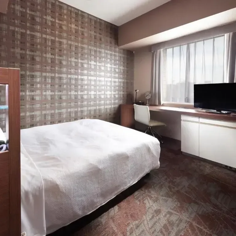 Modern hotel room with a neatly made white bed, work desk, chair, and television next to a window.