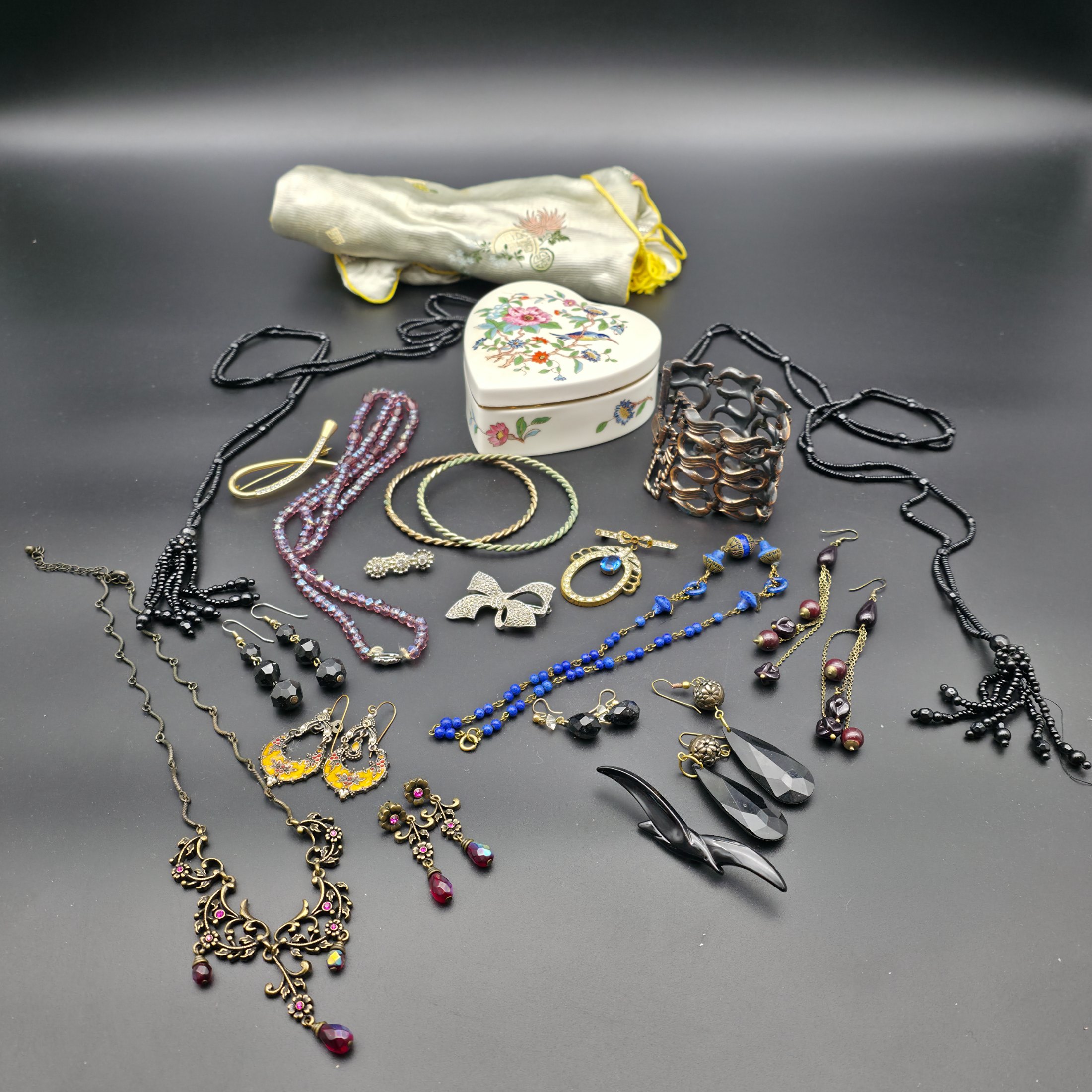 A diverse lot of vintage costume jewellery and decorative items from various eras, including necklaces, earrings, brooches, bracelets, a heart-shaped ceramic box, and a fabric pouch.