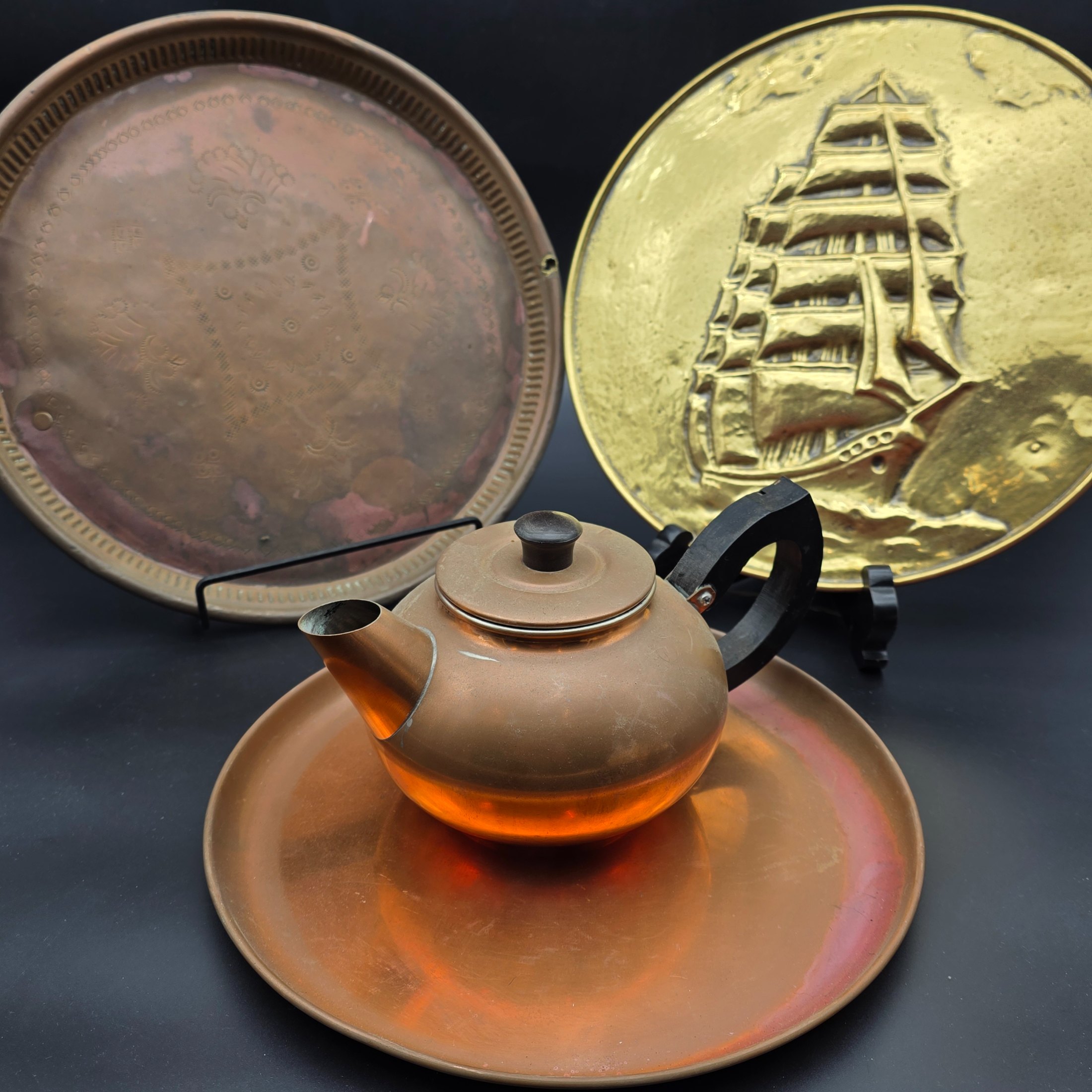 A collection of vintage copper and brass decorative and functional items, comprising two copper trays, a copper teapot, and an embossed brass wall plate depicting a sailing ship.
