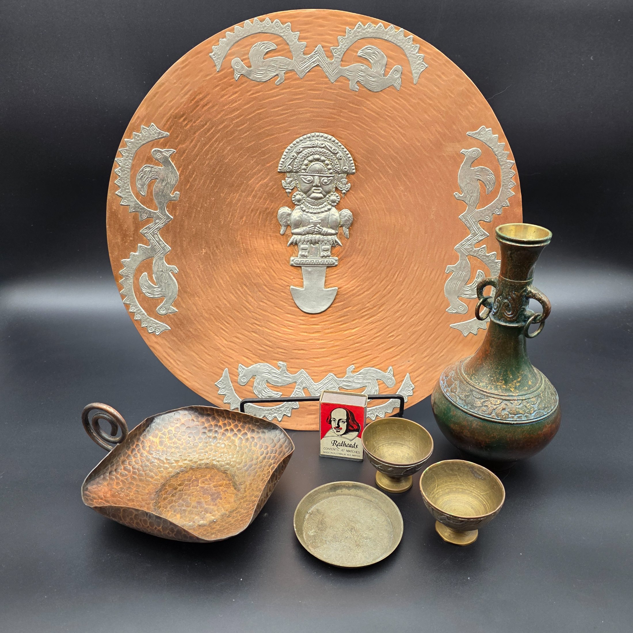 A diverse collection of vintage metalware, including a large copper wall plaque, a hammered copper dish, a small brass plate, two small brass cups, and a bronze/copper vase.
