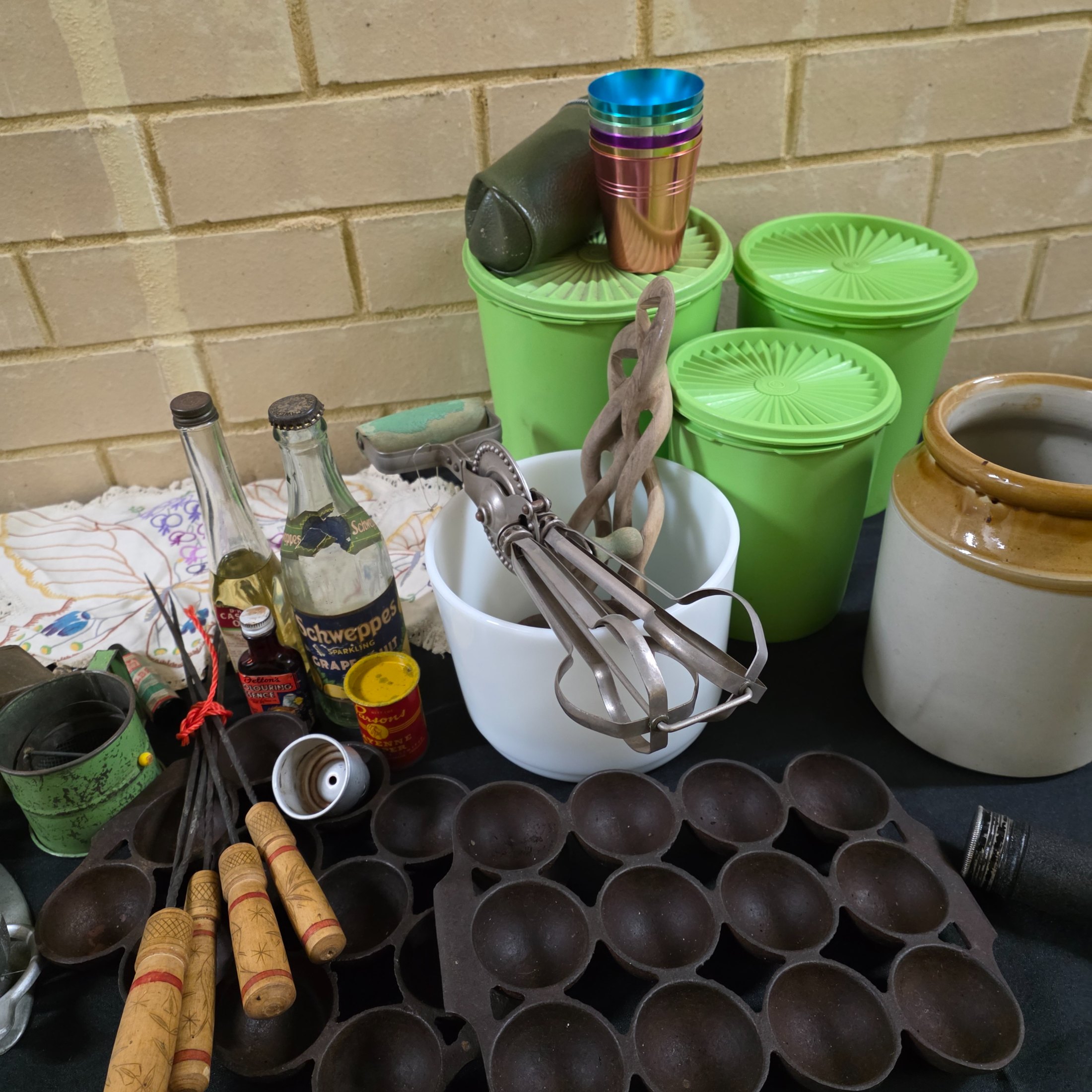 A diverse collection of vintage homewares and kitchen items including cast iron pans, a hand mixer, Tupperware, ceramic crocks, aluminium cups, and various other utensils and decorative pieces.