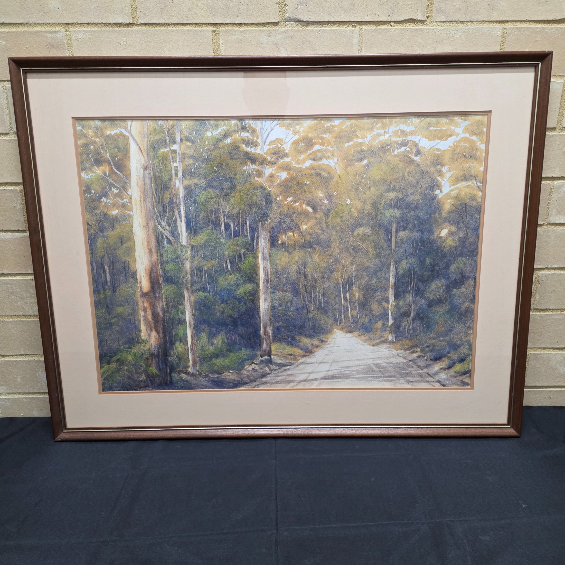 A framed original watercolour painting depicting a tranquil Australian bush scene with a dirt road winding through tall eucalypt trees, signed 'N. A. H.' and dated 1984.