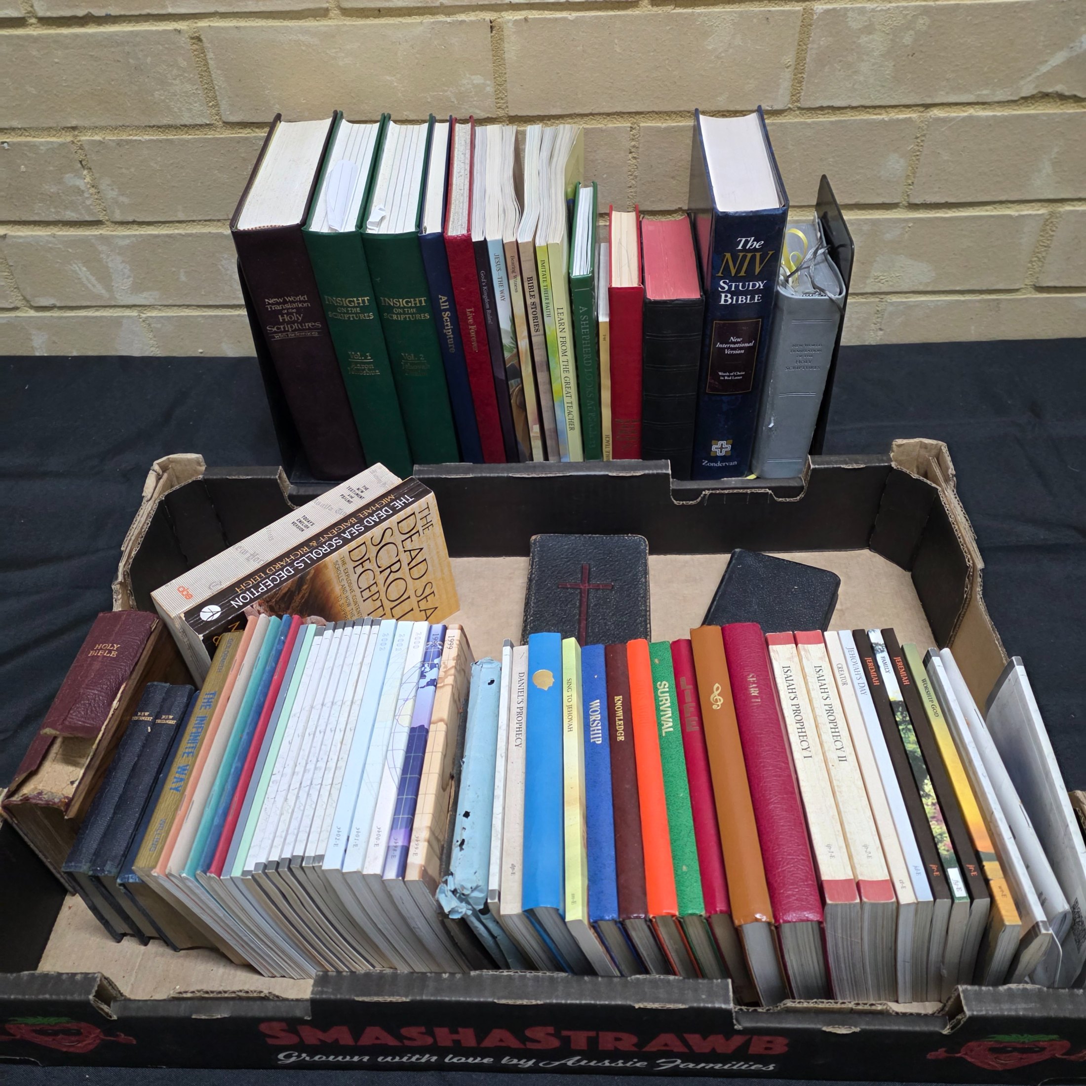 A large collection of mixed religious books and Bibles, including various translations, study guides, and annual publications, presented in a cardboard box and arranged on a surface.