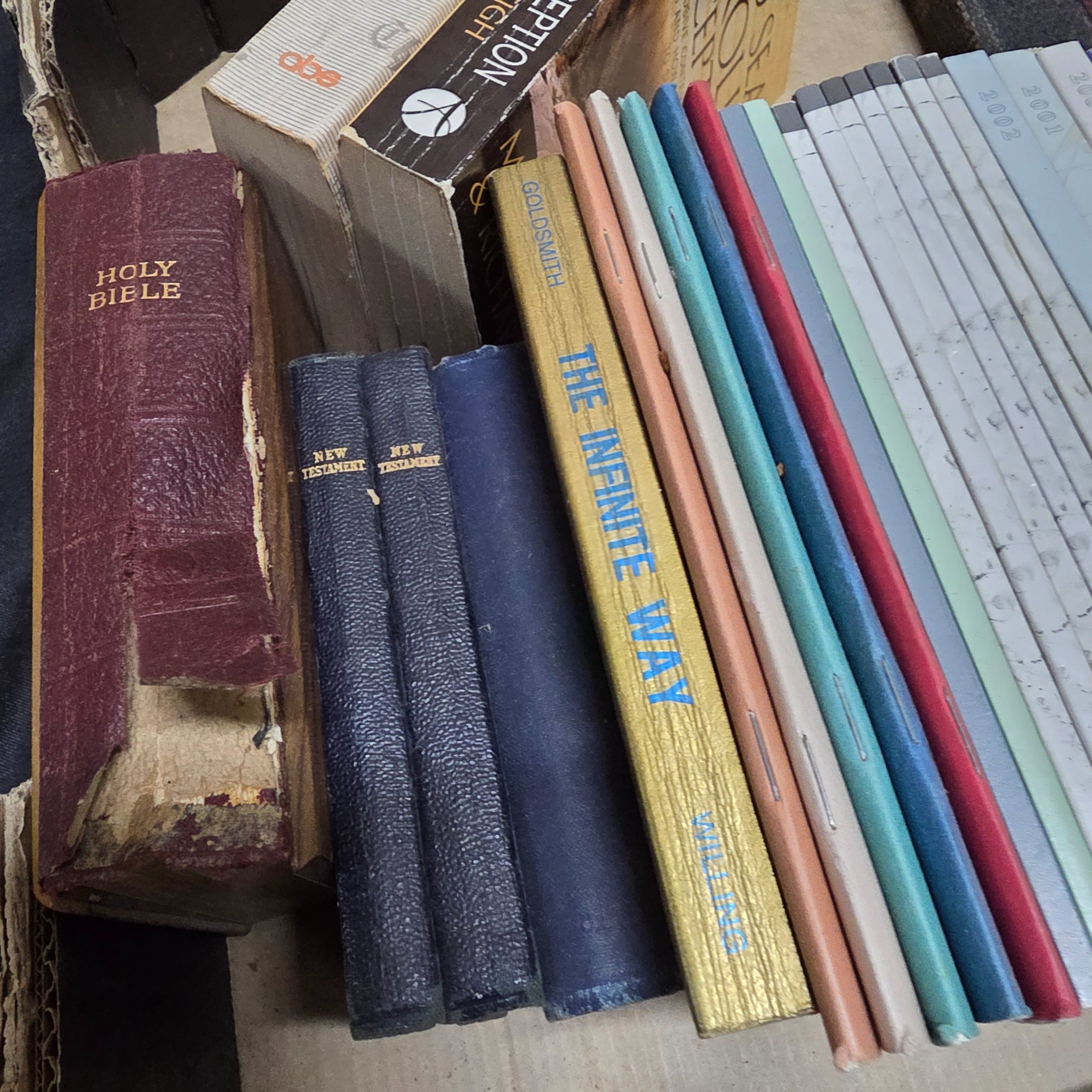 A large collection of mixed religious books and Bibles, including various translations, study guides, and annual publications, presented in a cardboard box and arranged on a surface. - View 7