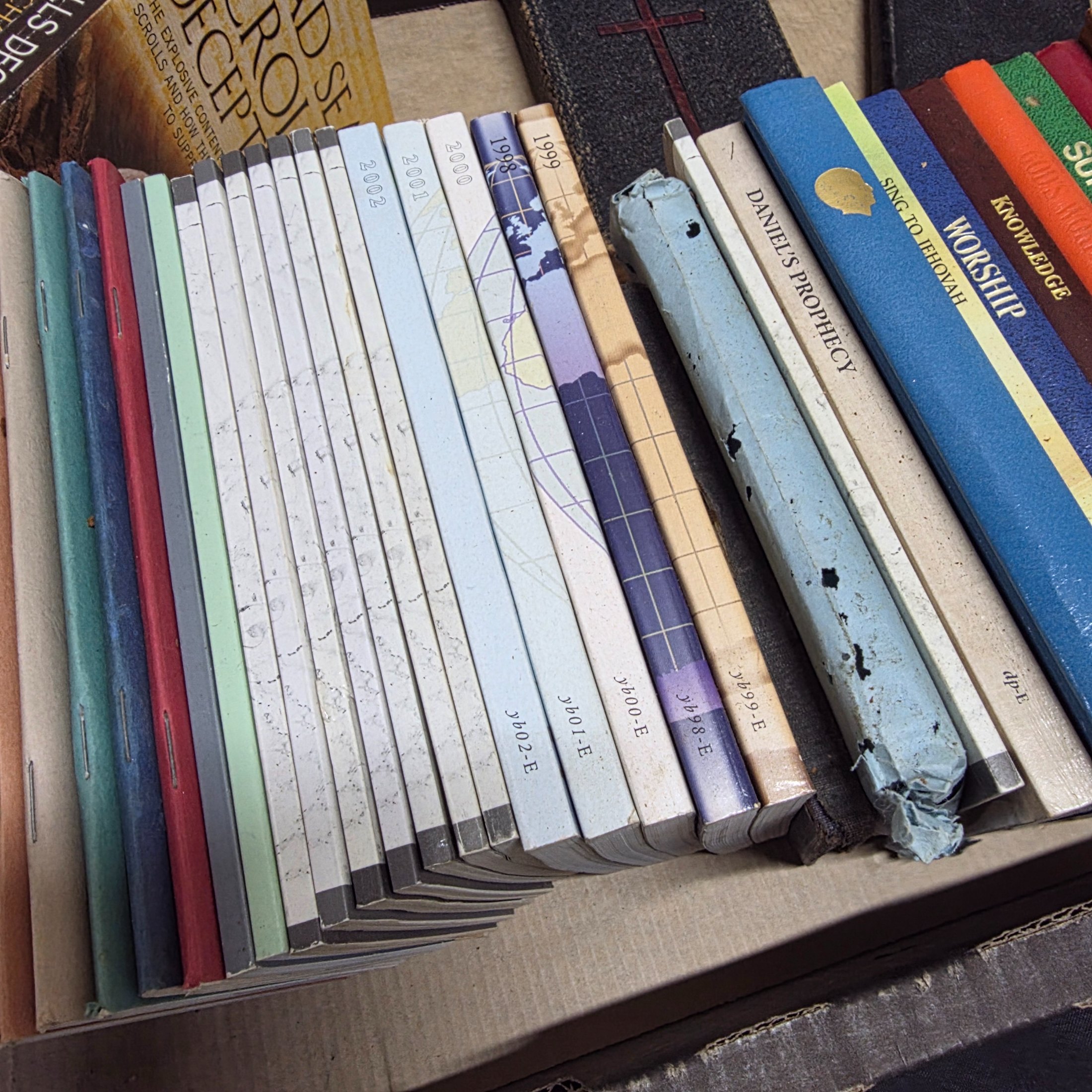 A large collection of mixed religious books and Bibles, including various translations, study guides, and annual publications, presented in a cardboard box and arranged on a surface. - View 8