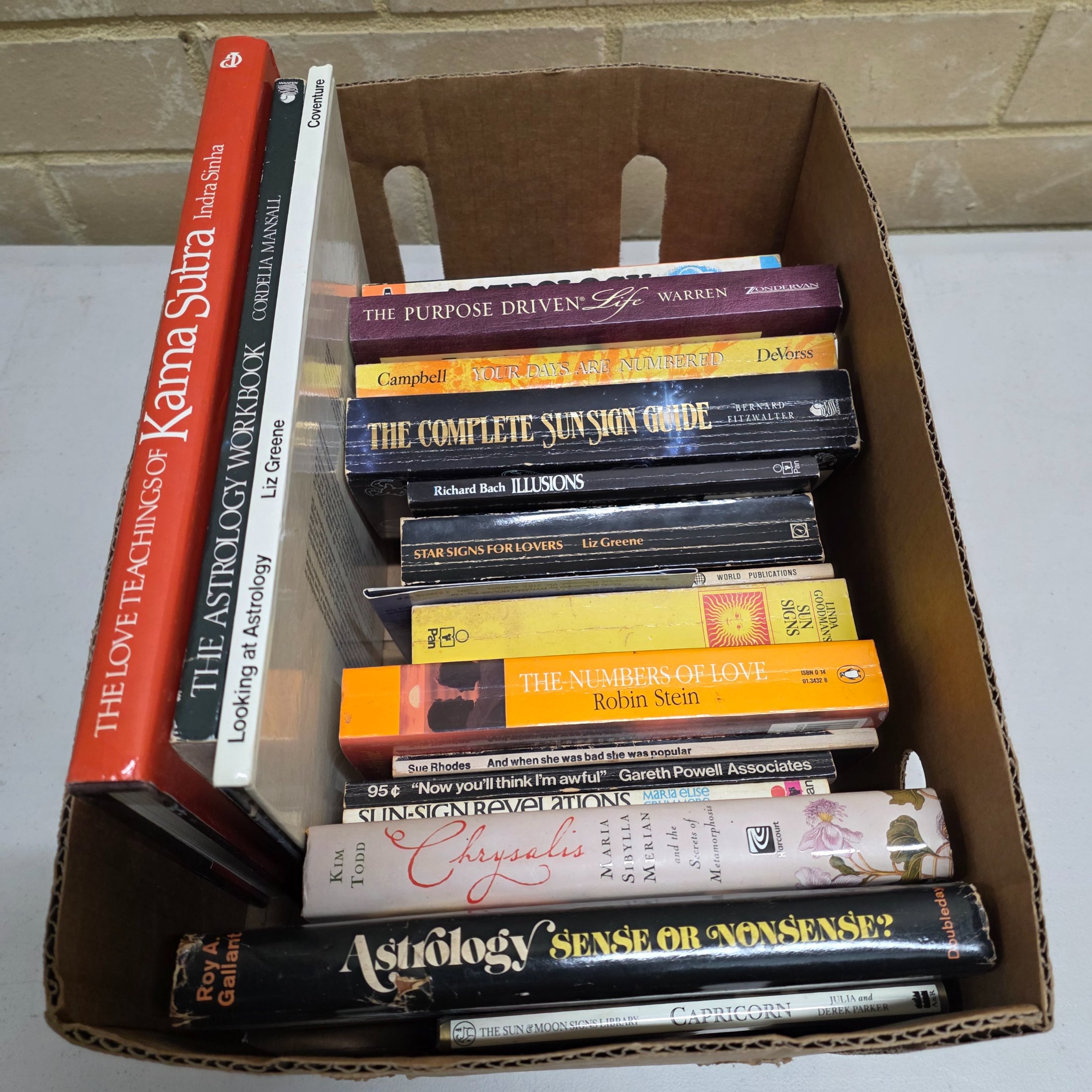 A cardboard box filled with approximately two dozen used books, predominantly on astrology, numerology, and self-help topics.