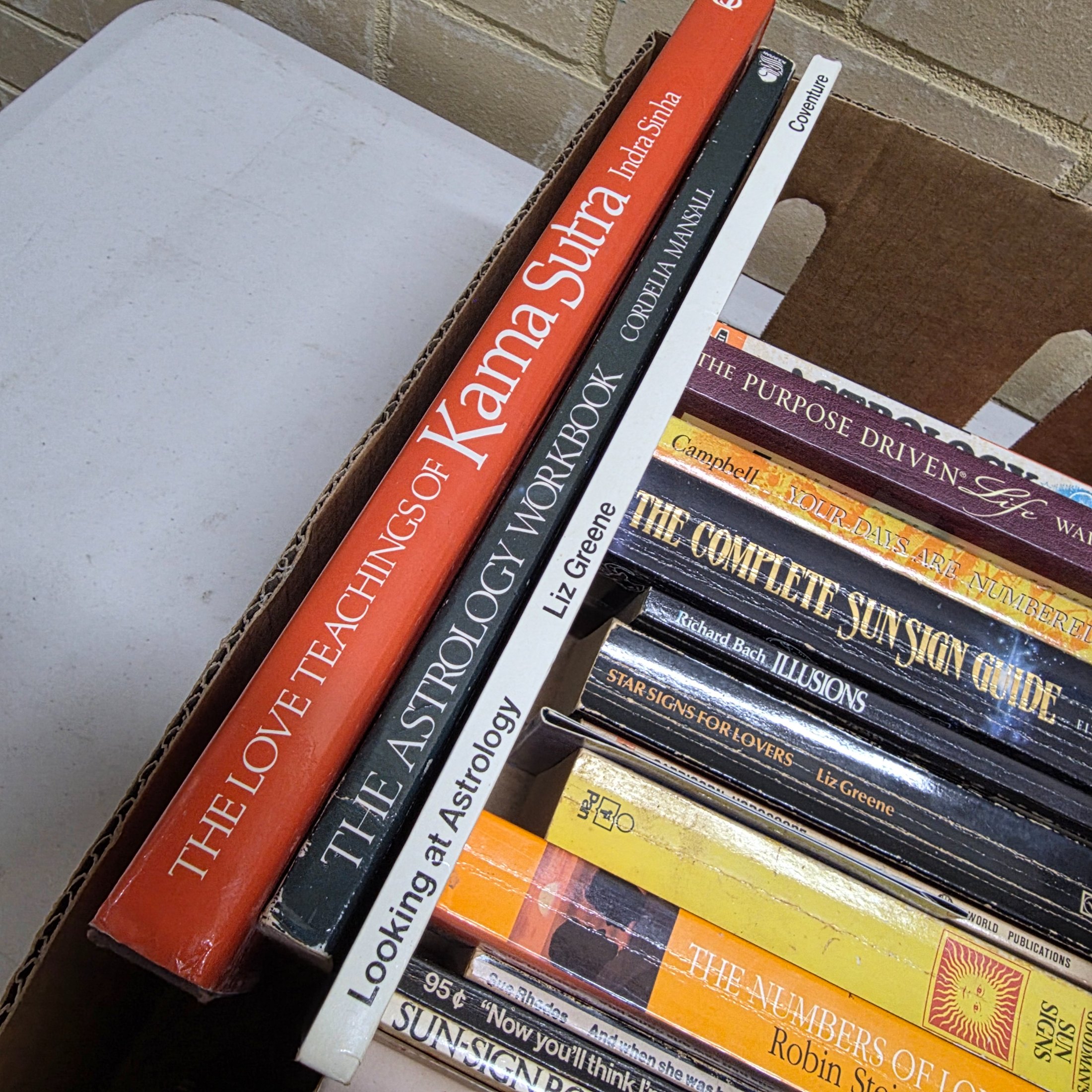 A cardboard box filled with approximately two dozen used books, predominantly on astrology, numerology, and self-help topics. - View 2