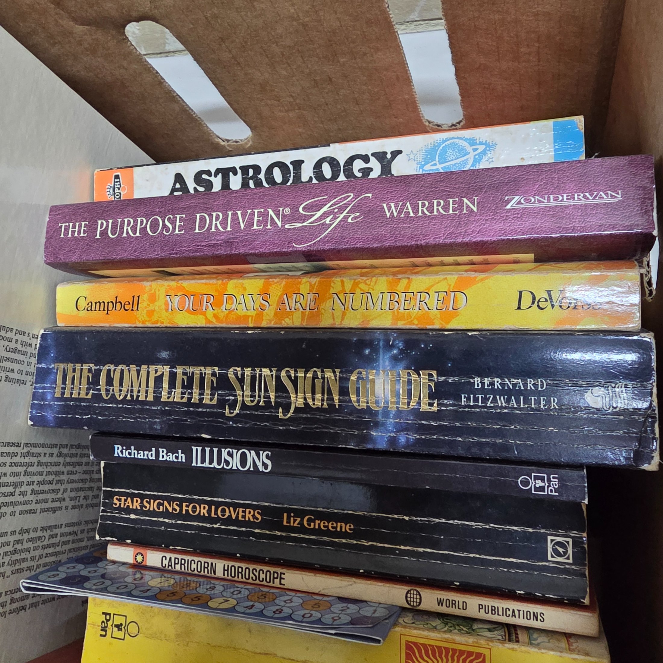 A cardboard box filled with approximately two dozen used books, predominantly on astrology, numerology, and self-help topics. - View 3