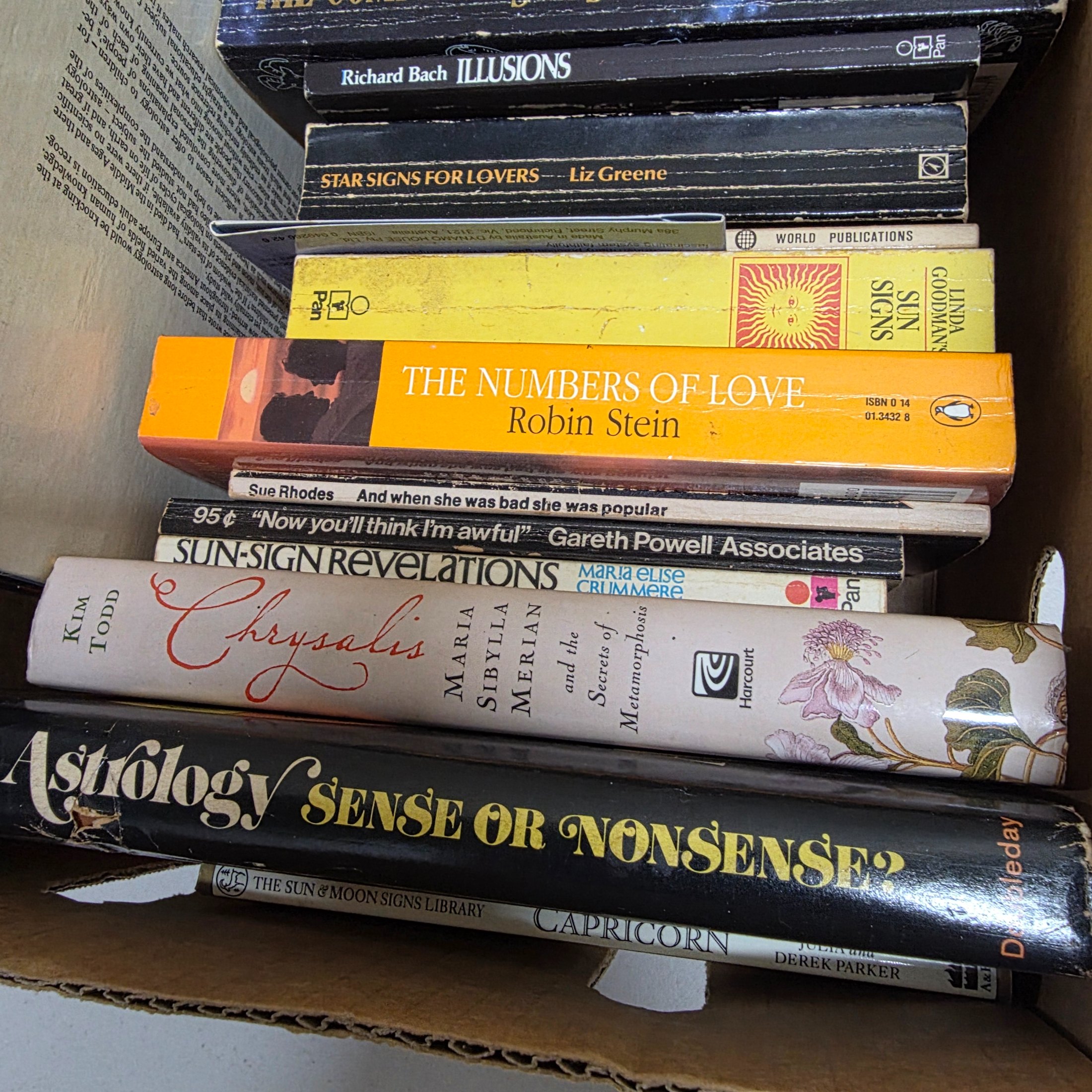 A cardboard box filled with approximately two dozen used books, predominantly on astrology, numerology, and self-help topics. - View 5