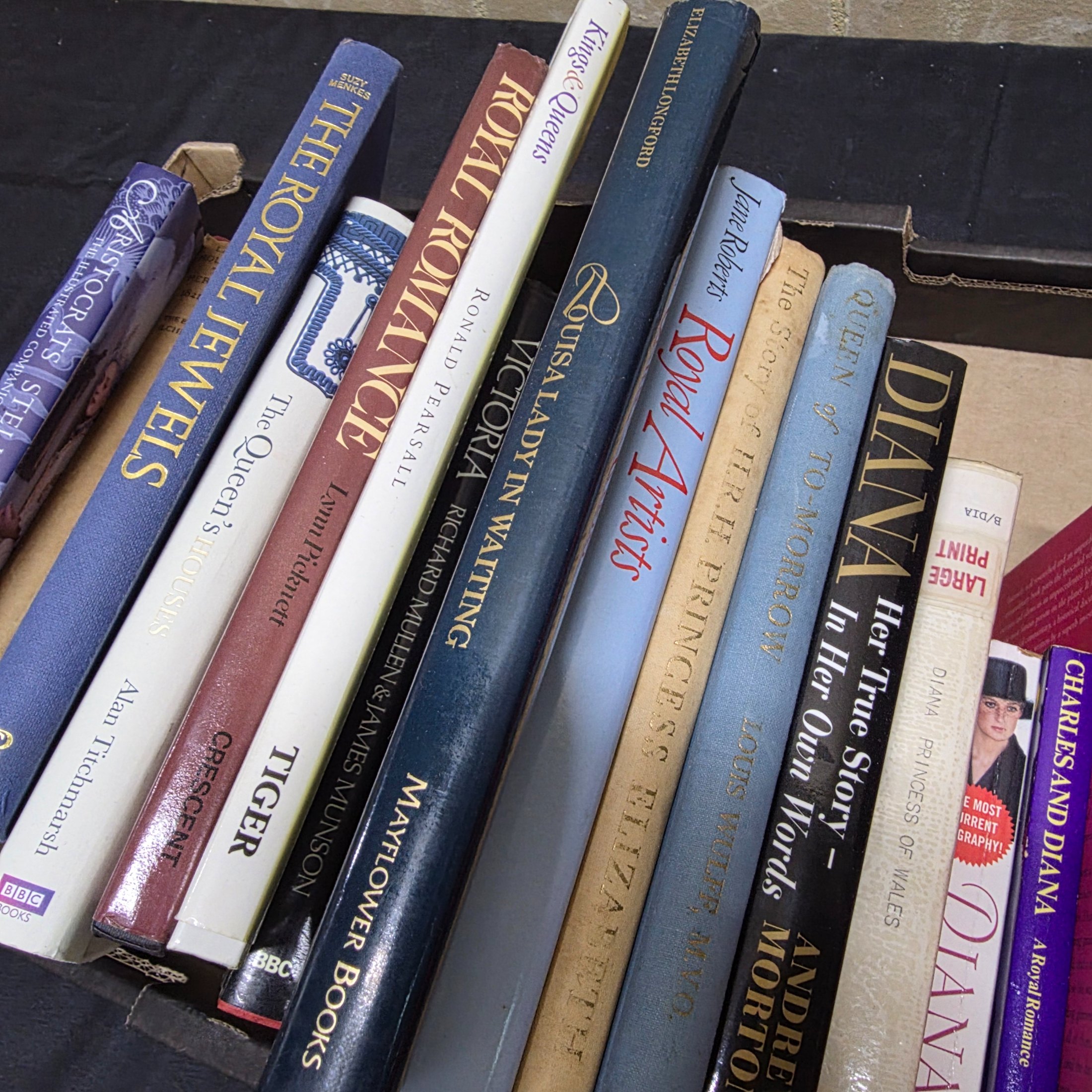 A cardboard box filled with approximately two dozen assorted books primarily focused on British royalty, including biographies of Princess Diana and historical accounts of castles and queens. - View 3