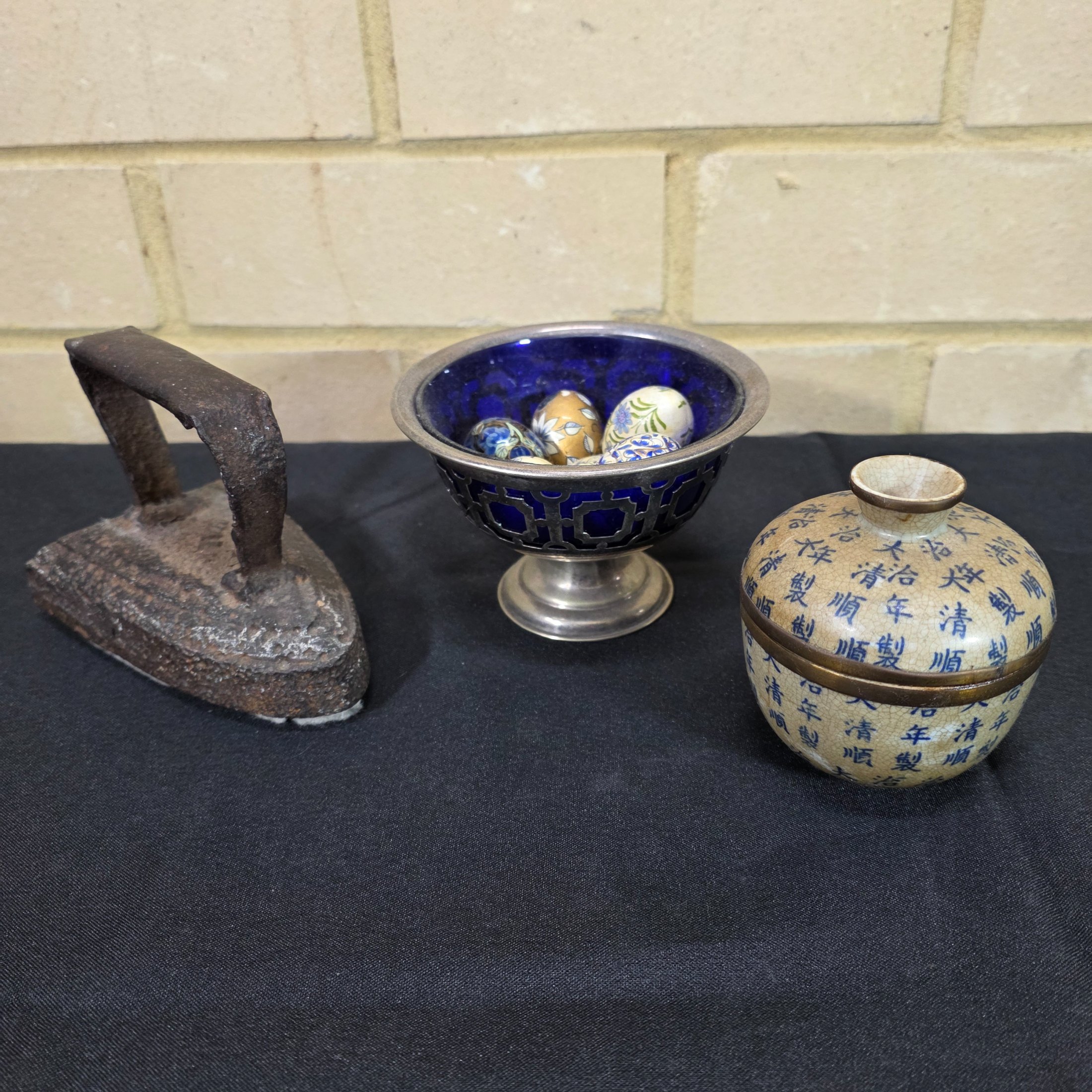 A diverse lot of antique and vintage items, including a rusty cast iron flat iron, a silver-plated bowl with a cobalt blue glass insert holding decorative painted eggs, and a Chinese crackle-glazed ceramic lidded jar with blue characters.