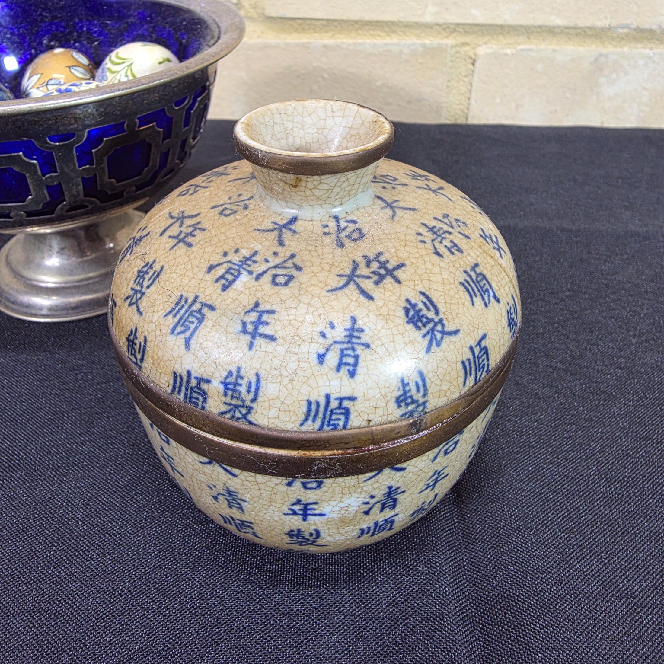 A diverse lot of antique and vintage items, including a rusty cast iron flat iron, a silver-plated bowl with a cobalt blue glass insert holding decorative painted eggs, and a Chinese crackle-glazed ceramic lidded jar with blue characters. - View 2