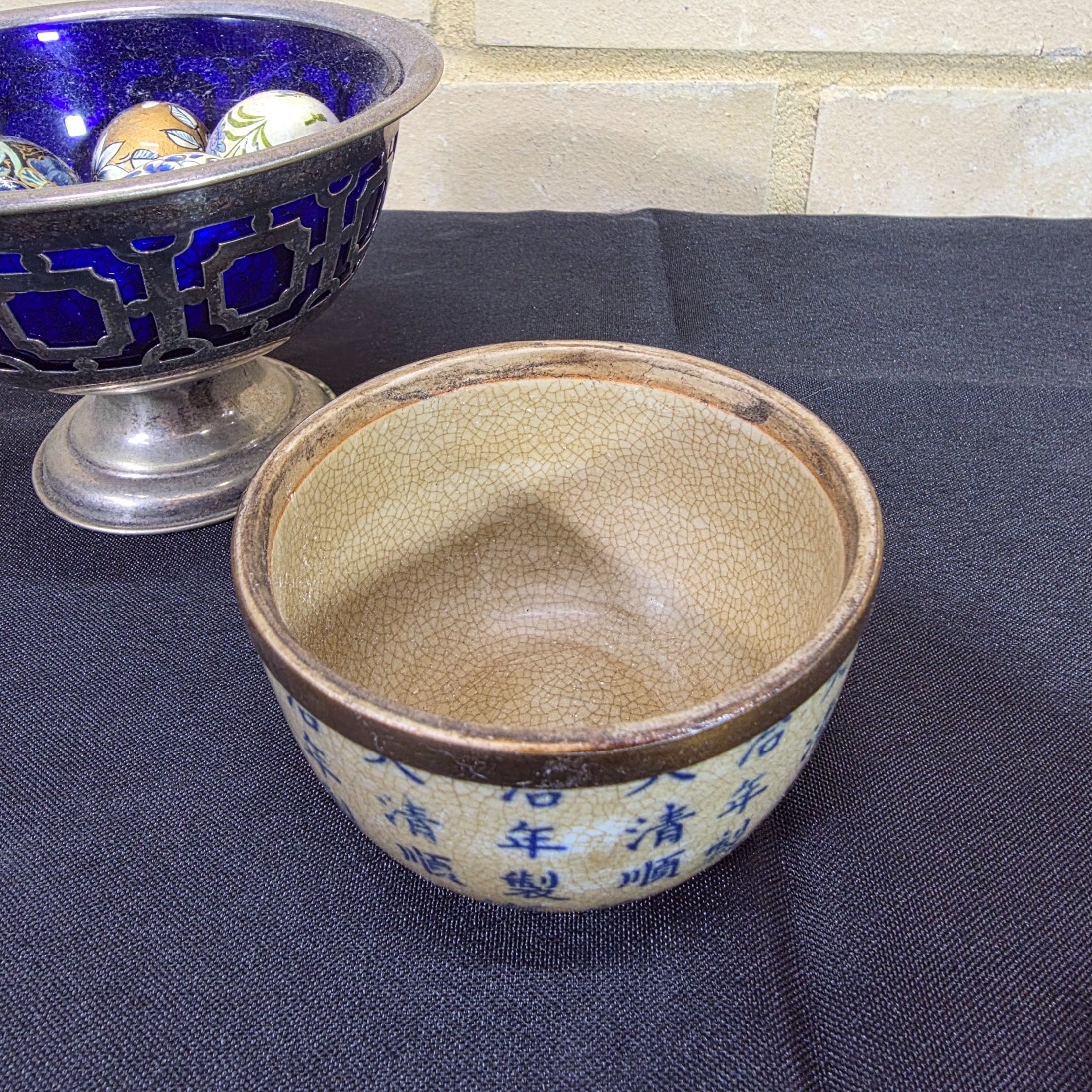 A diverse lot of antique and vintage items, including a rusty cast iron flat iron, a silver-plated bowl with a cobalt blue glass insert holding decorative painted eggs, and a Chinese crackle-glazed ceramic lidded jar with blue characters. - View 3