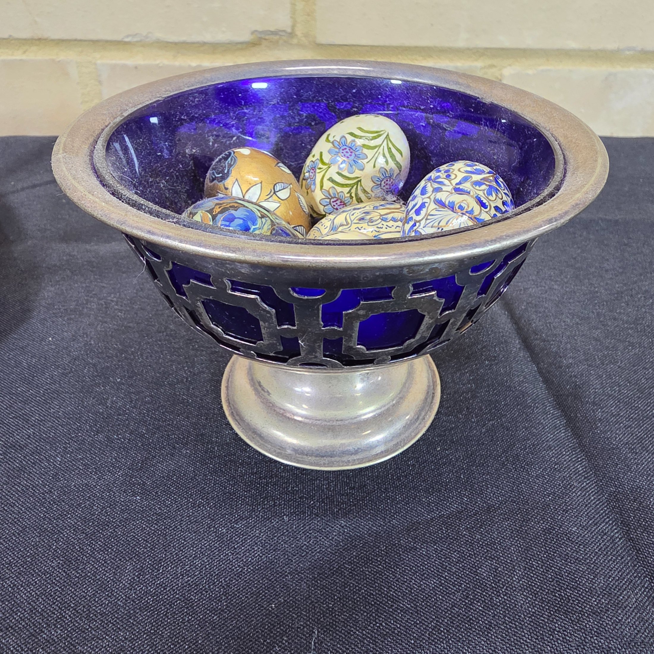 A diverse lot of antique and vintage items, including a rusty cast iron flat iron, a silver-plated bowl with a cobalt blue glass insert holding decorative painted eggs, and a Chinese crackle-glazed ceramic lidded jar with blue characters. - View 7