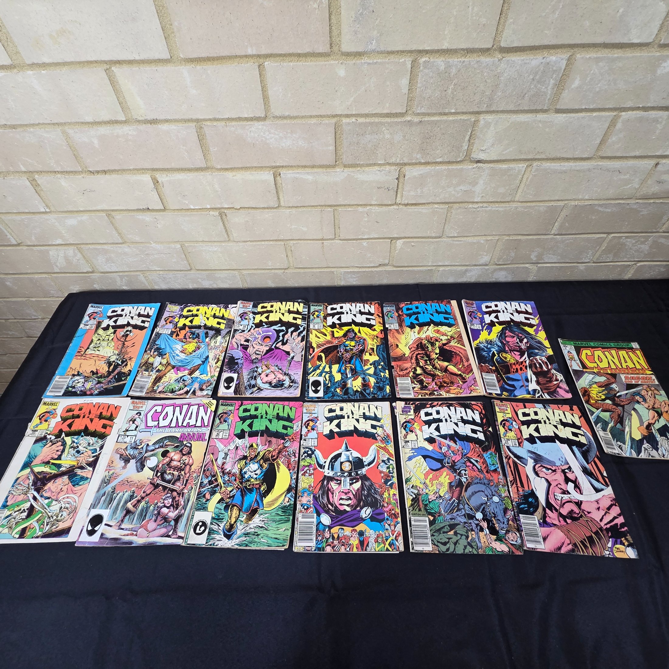 A collection of vintage Marvel 'Conan the King' and 'Conan the Barbarian' comic books from the 1980s, displayed on a black surface against a brick wall.
