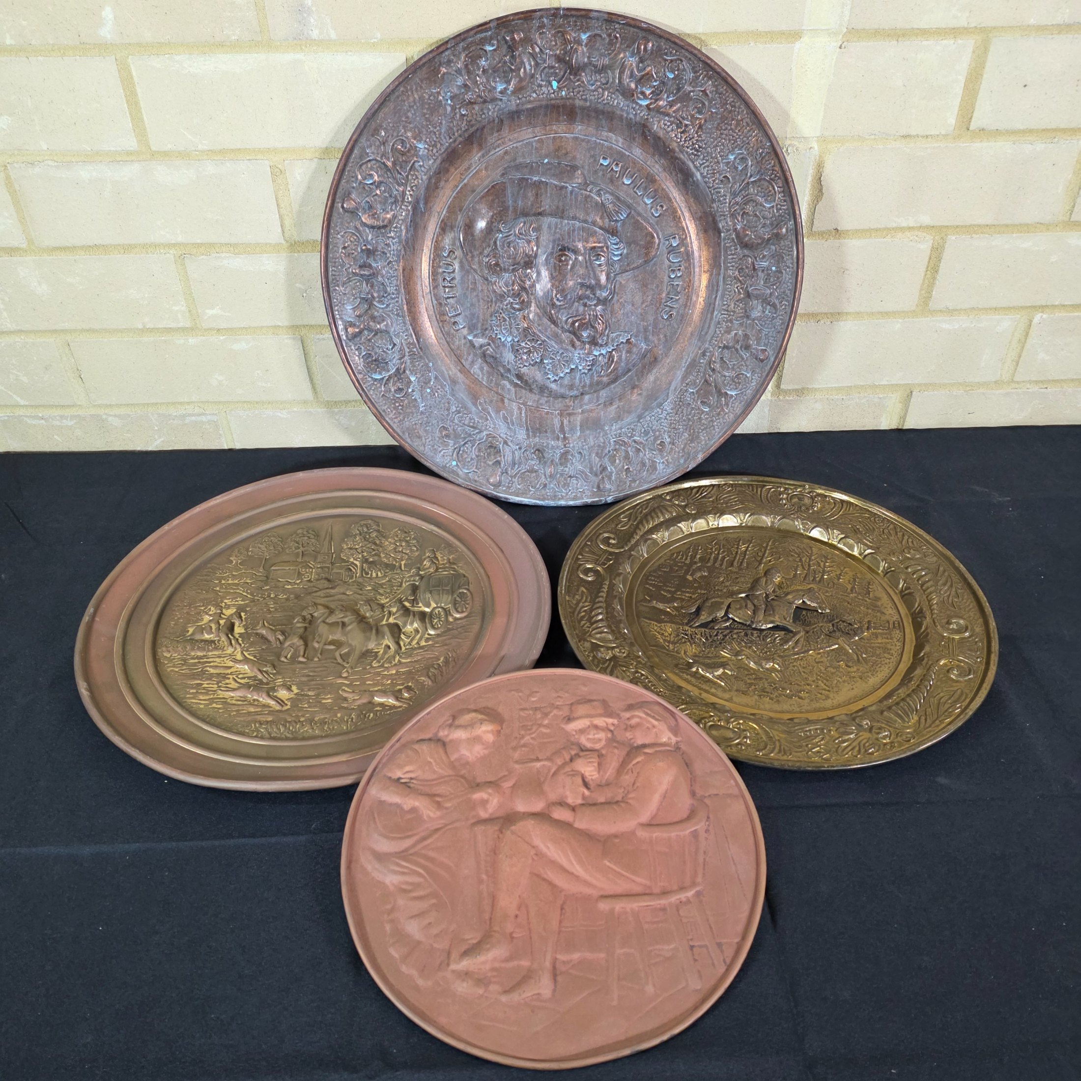 A collection of four vintage embossed metal wall plaques, comprising a large copper-toned plaque featuring a portrait of Peter Paul Rubens, two brass-toned plaques depicting fox hunting and horse-drawn carriage scenes, and a smaller copper-toned plaque with a tavern scene.