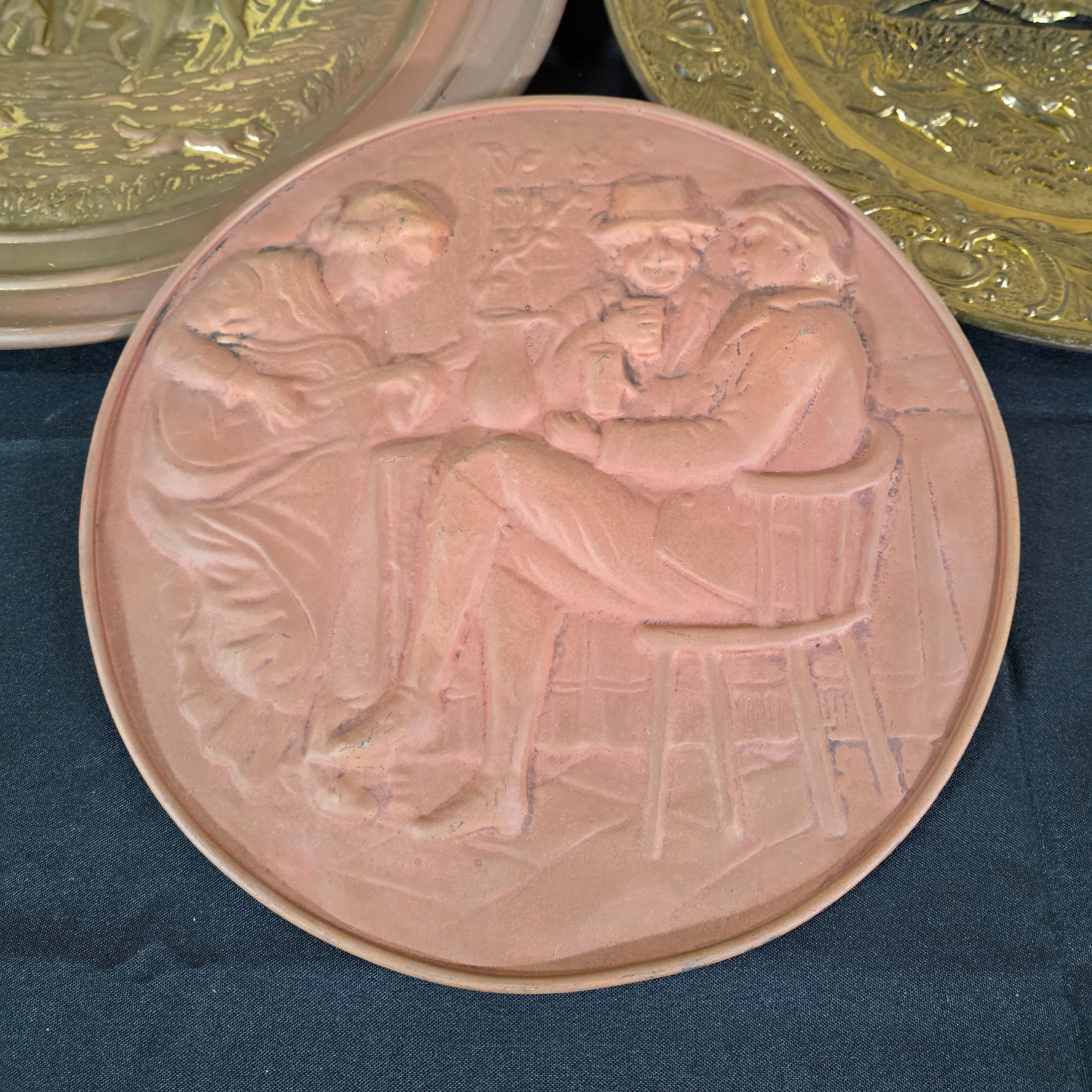 A collection of four vintage embossed metal wall plaques, comprising a large copper-toned plaque featuring a portrait of Peter Paul Rubens, two brass-toned plaques depicting fox hunting and horse-drawn carriage scenes, and a smaller copper-toned plaque with a tavern scene. - View 7