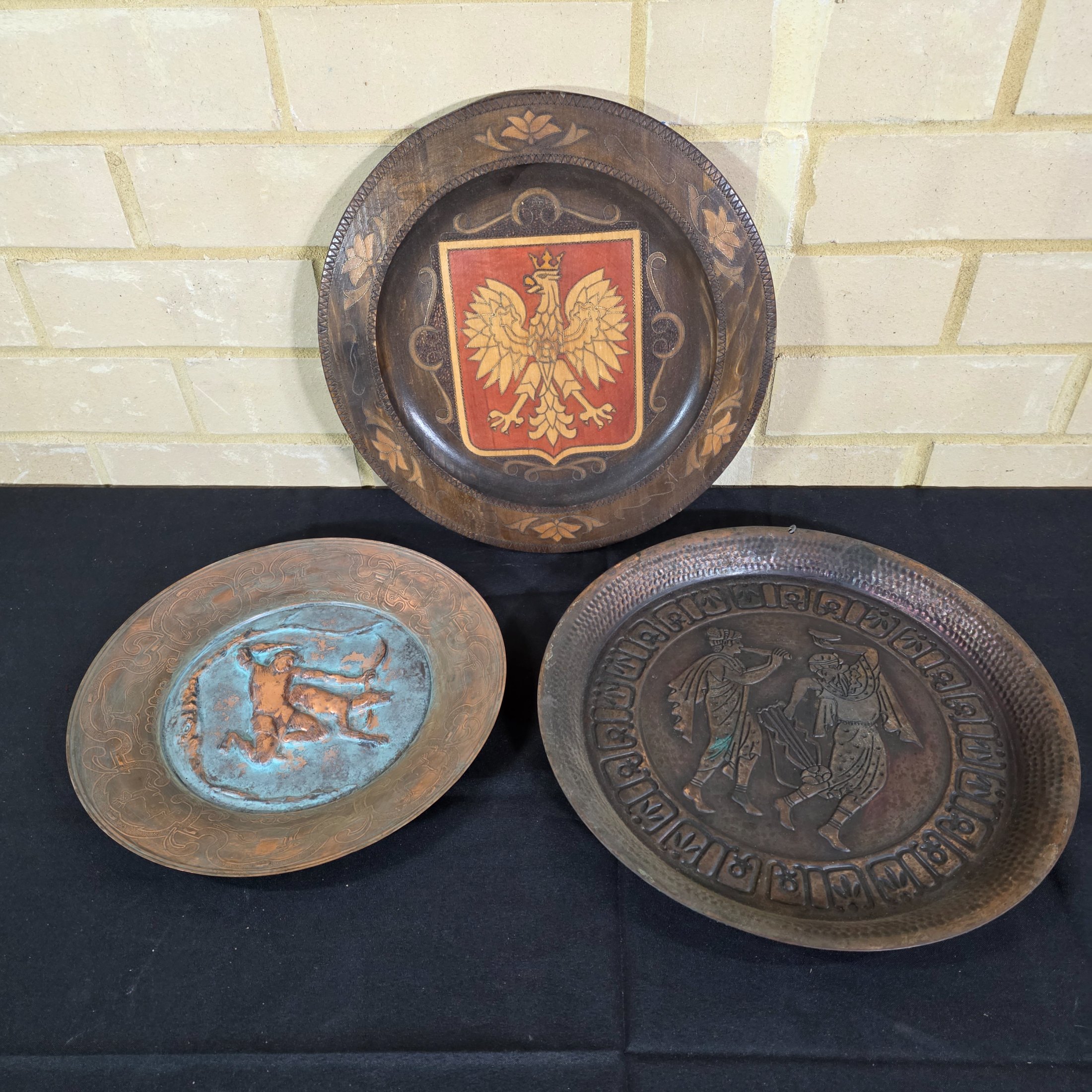A collection of three vintage decorative wall plates, including a wooden plate with a Polish eagle crest and two copper or bronze plates featuring classical hunting and musical figures.