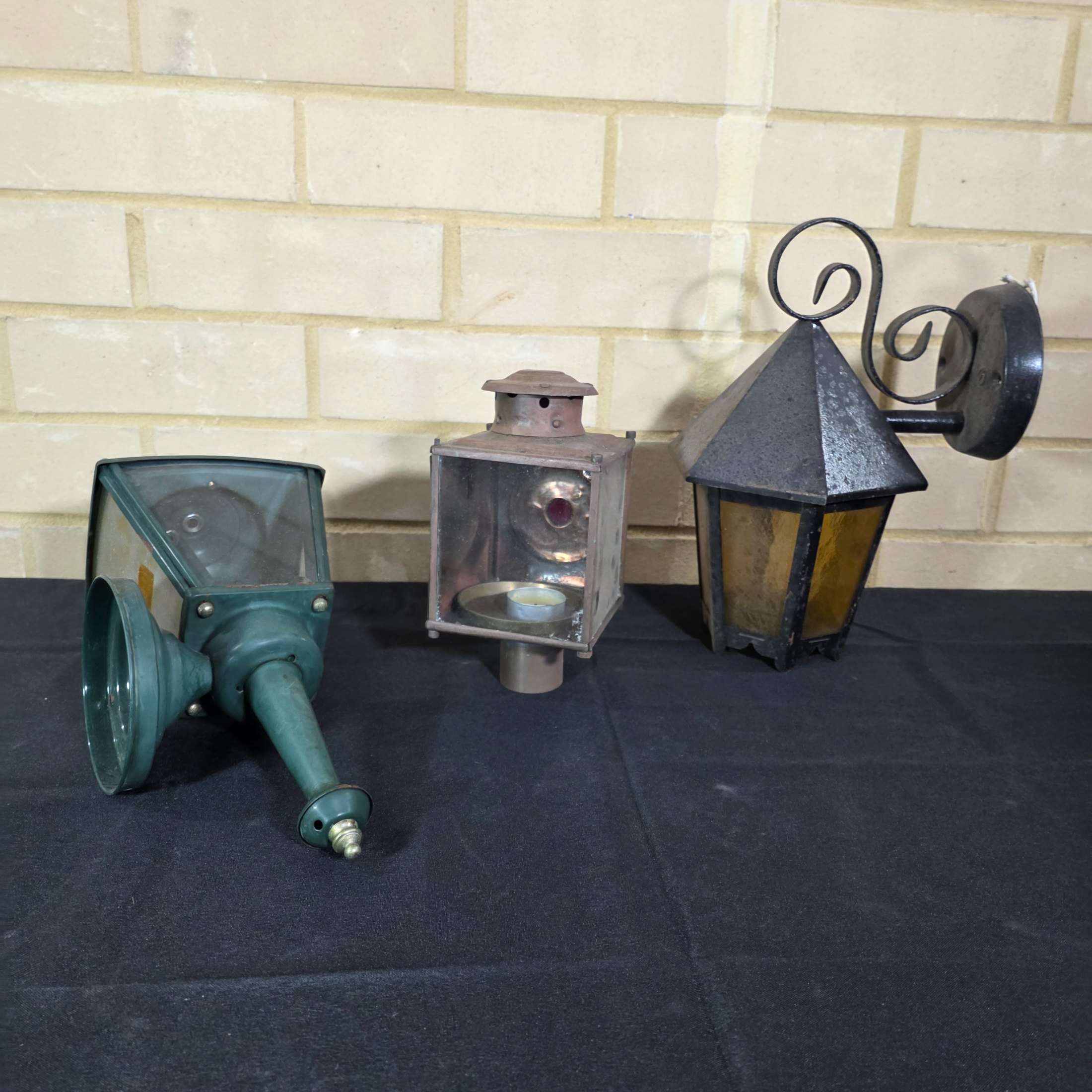 A diverse lot of three vintage outdoor lanterns, comprising a green wall-mounted electric light, an older copper-toned candle lantern, and a black painted Kempthorne electric wall sconce with amber glass.