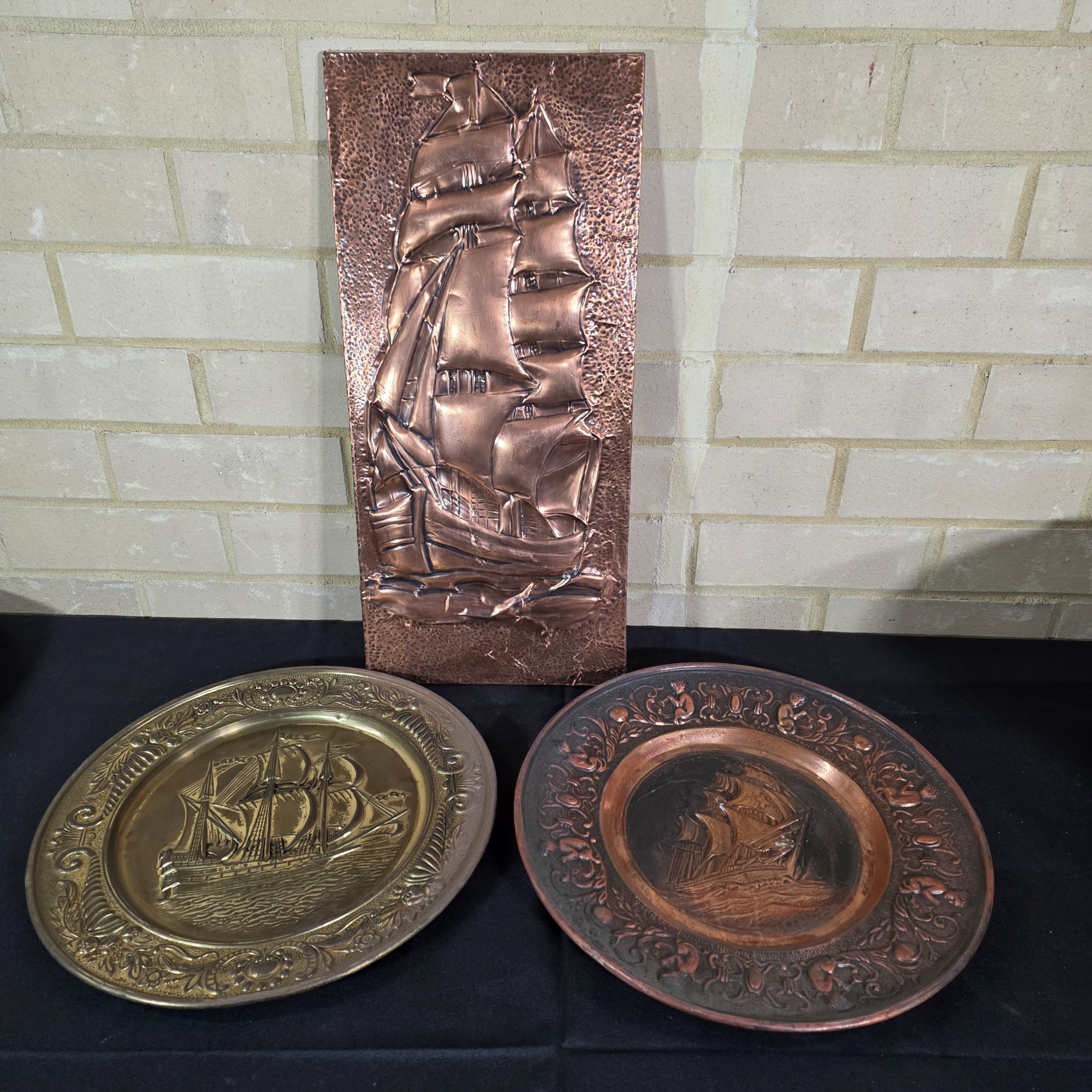A collection of three vintage embossed metal decorative items, including a rectangular copper wall hanging and two round plates, all depicting sailing ships.