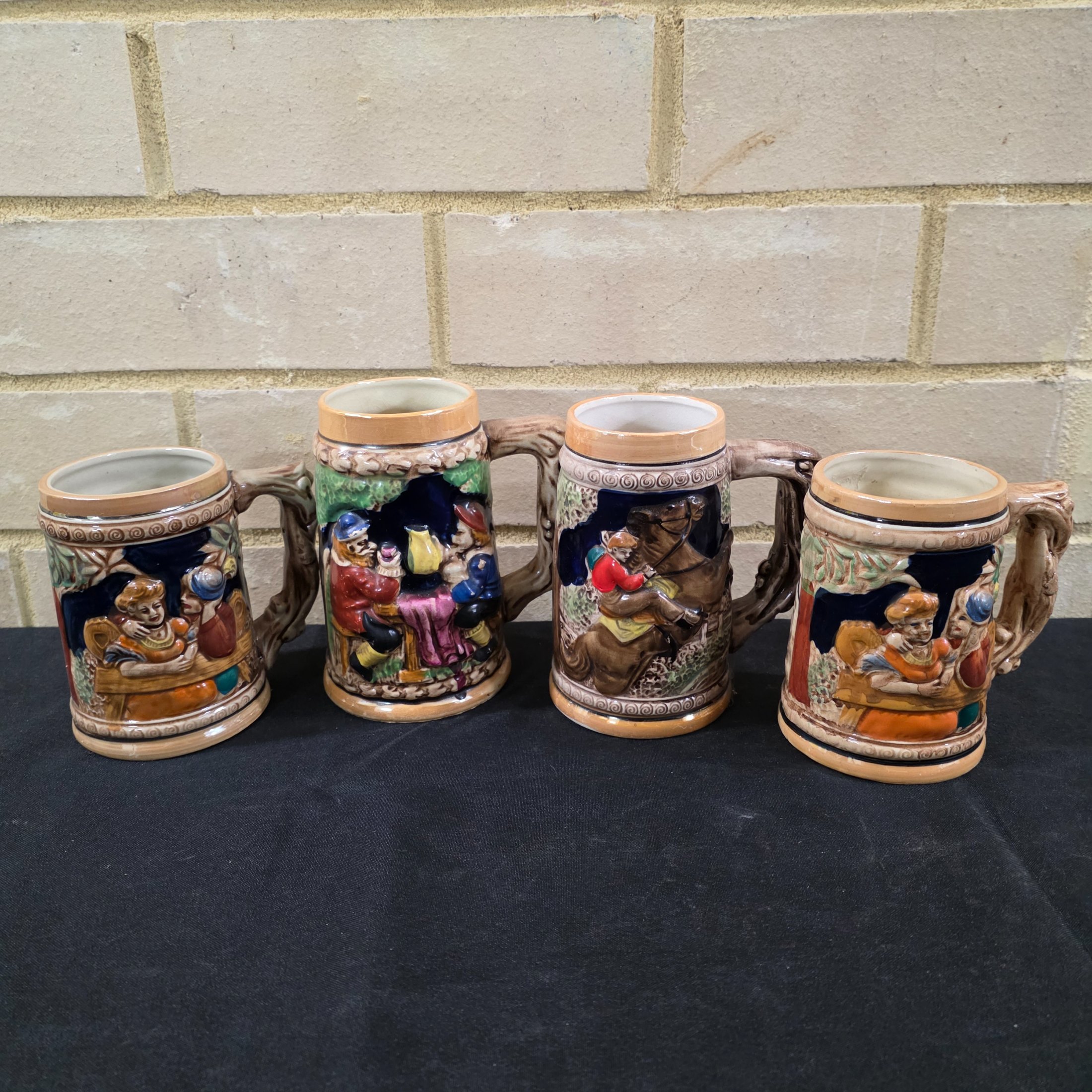 A set of four vintage Japanese ceramic mugs, designed in the style of traditional German beer steins, featuring intricate, hand-painted raised relief scenes depicting various Bavarian-themed activities and figures.