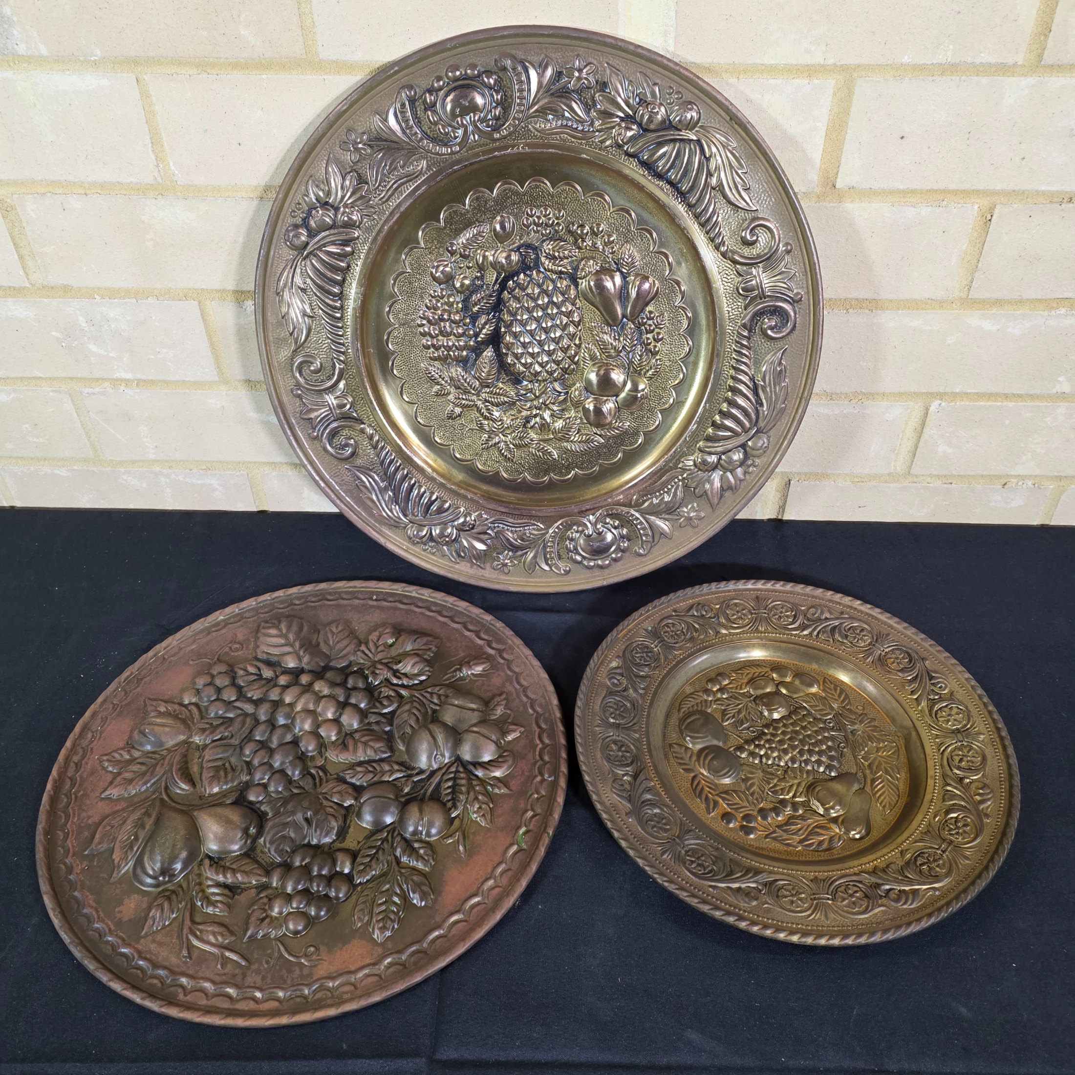 A set of three vintage decorative wall plaques, featuring embossed fruit designs in brass and copper-toned metal.