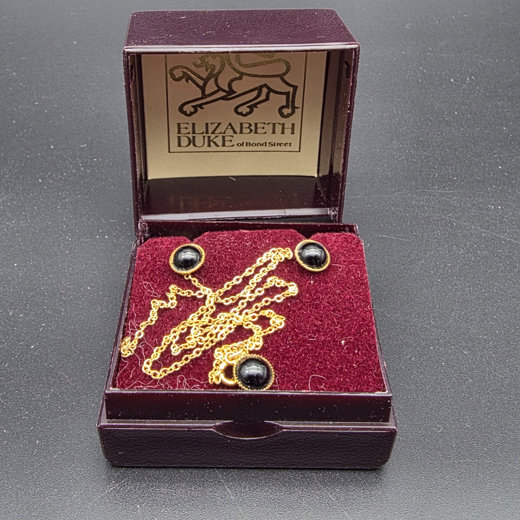 A vintage Elizabeth Duke of Bond Street jewellery set, crafted from 9ct gold and featuring elegant black cabochon stones, presented in its original branded box. This classic ensemble comprises a delicate chain necklace and matching stud earrings.