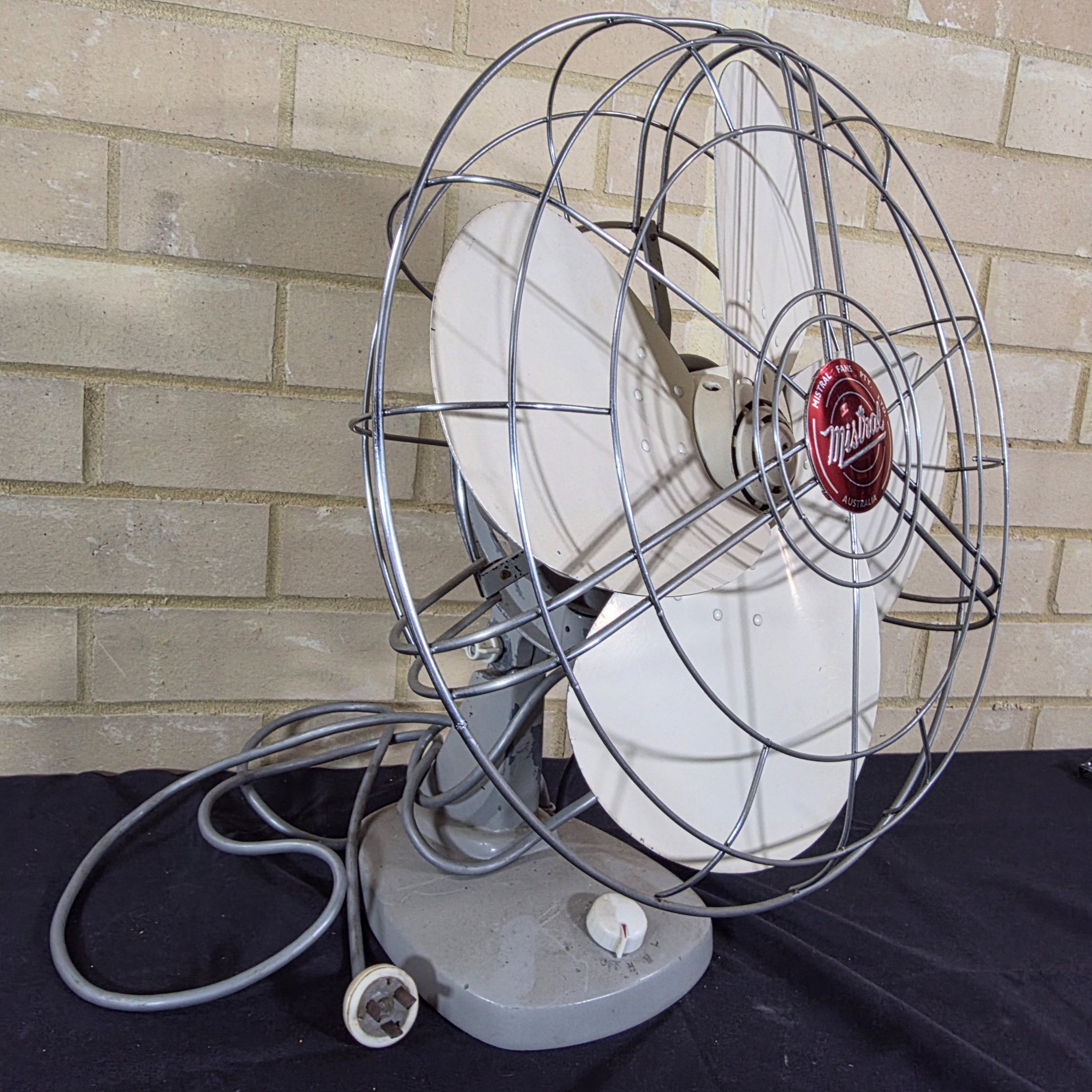 A vintage Mistral electric fan, likely from the mid-20th century, featuring a grey painted metal base and motor housing, a chrome wire cage, and cream plastic blades, with a visible red Mistral logo on the front grille.
