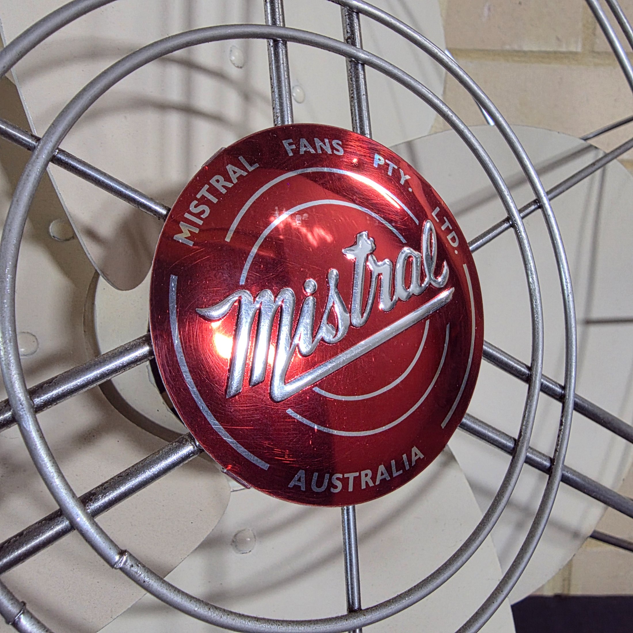 A vintage Mistral electric fan, likely from the mid-20th century, featuring a grey painted metal base and motor housing, a chrome wire cage, and cream plastic blades, with a visible red Mistral logo on the front grille. - View 2