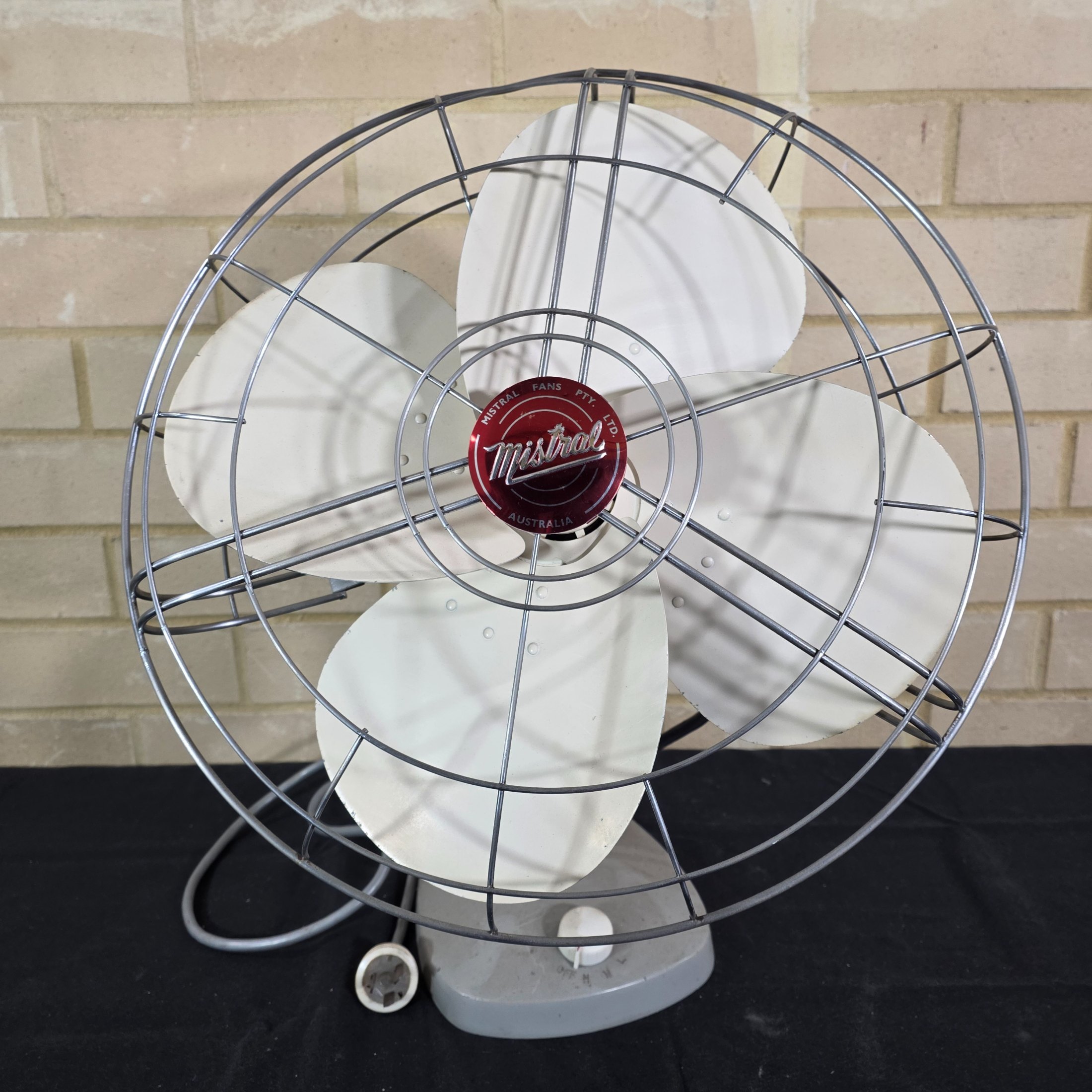 A vintage Mistral electric fan, likely from the mid-20th century, featuring a grey painted metal base and motor housing, a chrome wire cage, and cream plastic blades, with a visible red Mistral logo on the front grille. - View 3