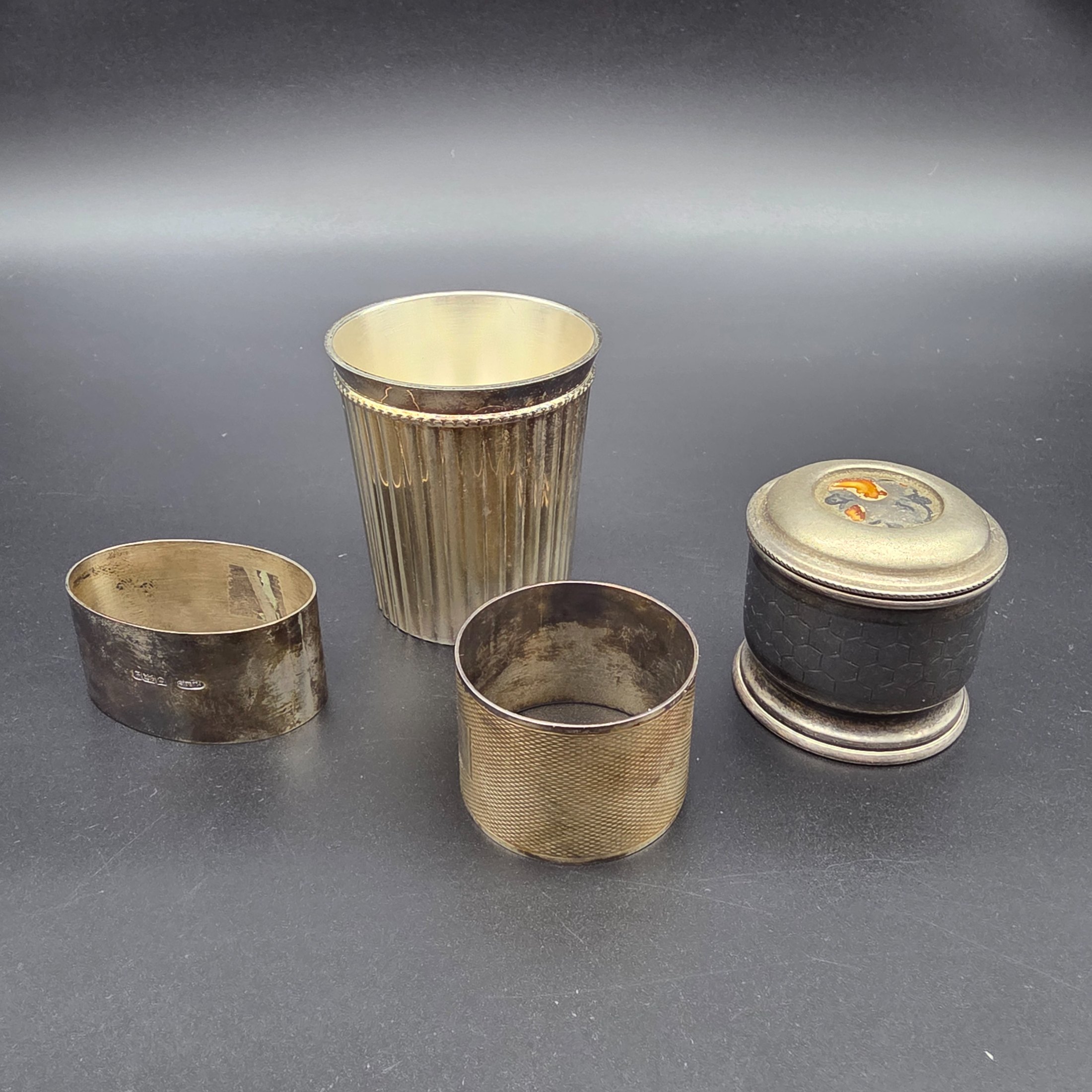 A collection of four vintage silver-toned items, including a sterling silver napkin ring, a second napkin ring, a ribbed cup, and a lidded box, dating from the early to mid-20th century.
