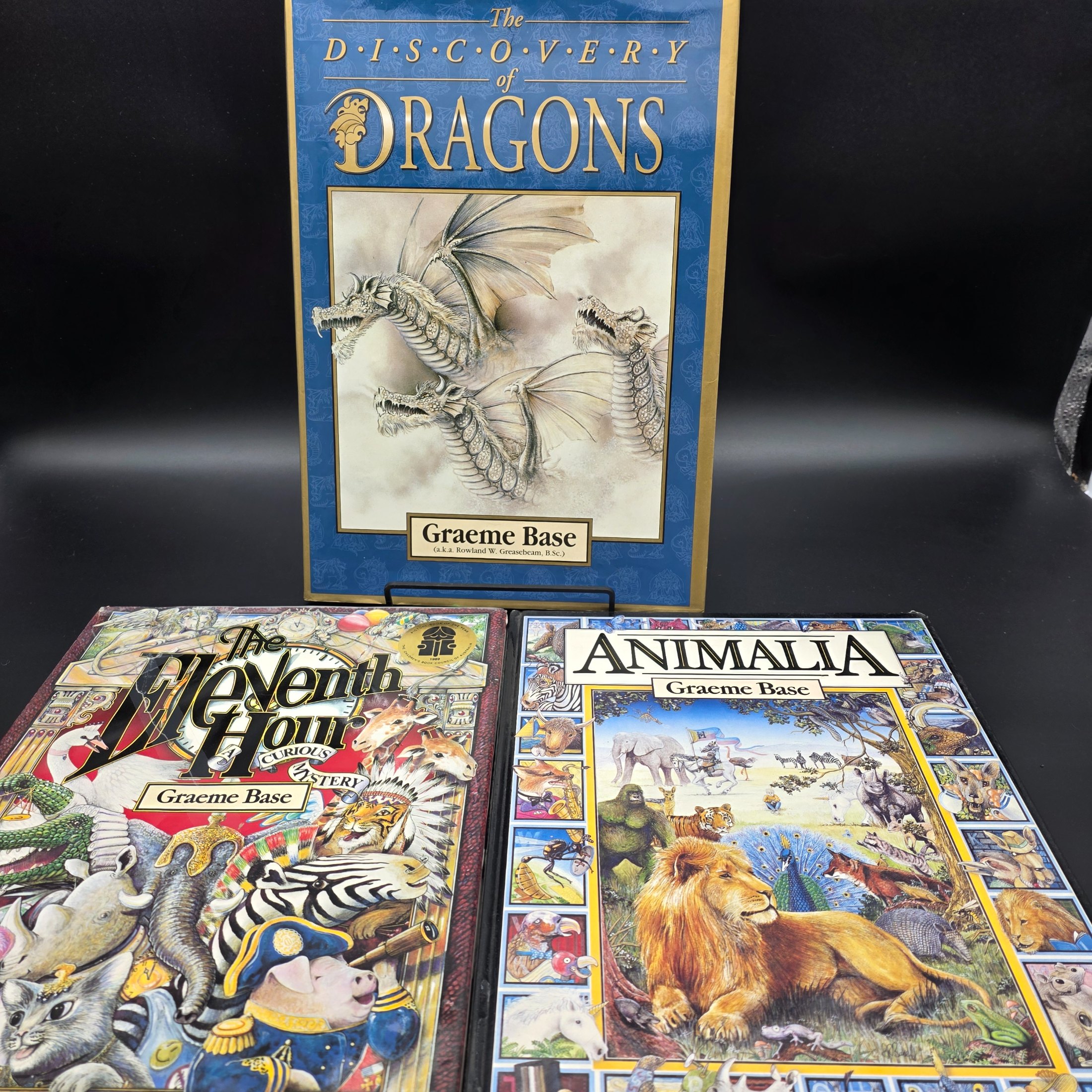 A collection of three hardcover children's books by Graeme Base: 'The Discovery of Dragons', 'The Eleventh Hour Curious Mystery', and 'Animalia', featuring intricate illustrations.