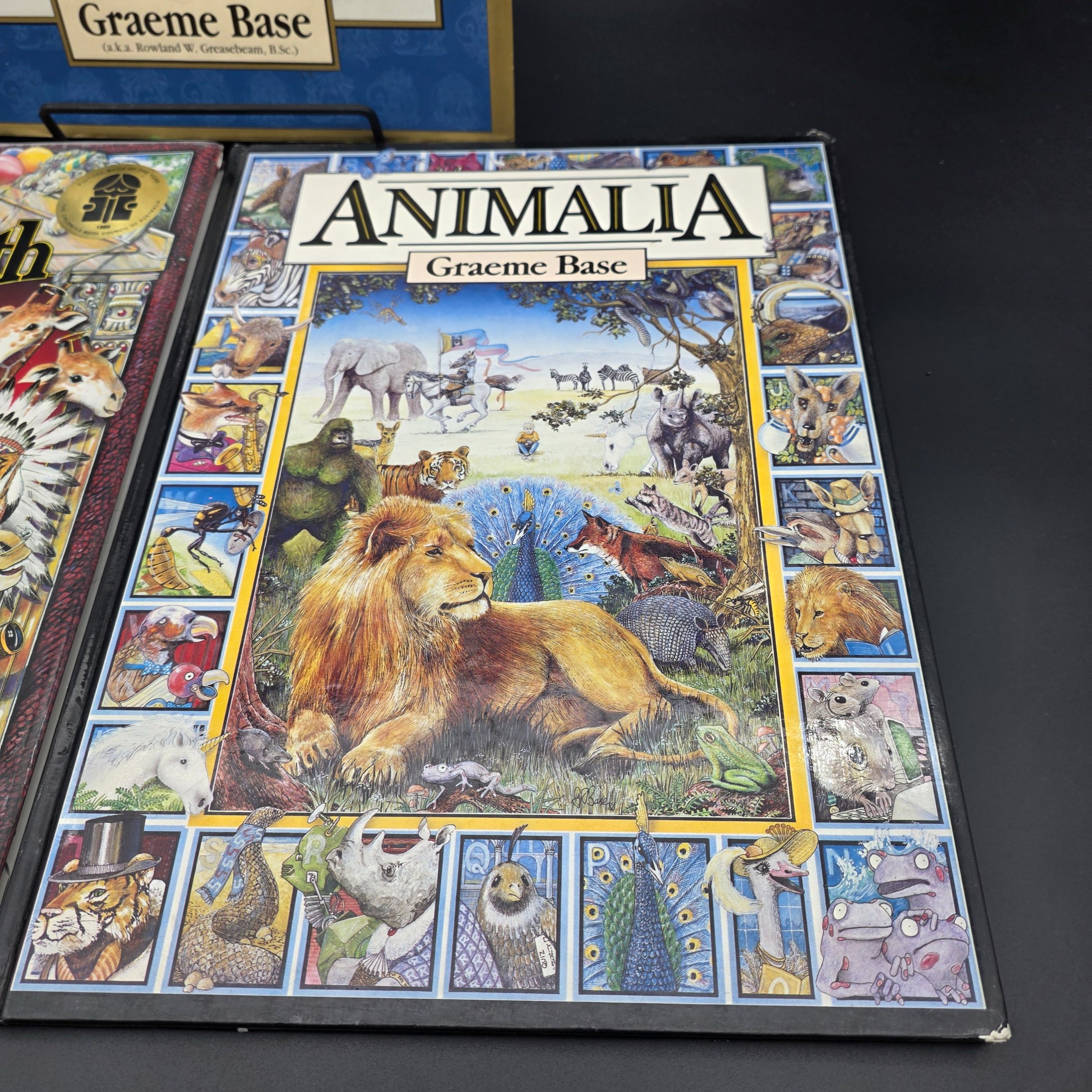 A collection of three hardcover children's books by Graeme Base: 'The Discovery of Dragons', 'The Eleventh Hour Curious Mystery', and 'Animalia', featuring intricate illustrations. - View 2