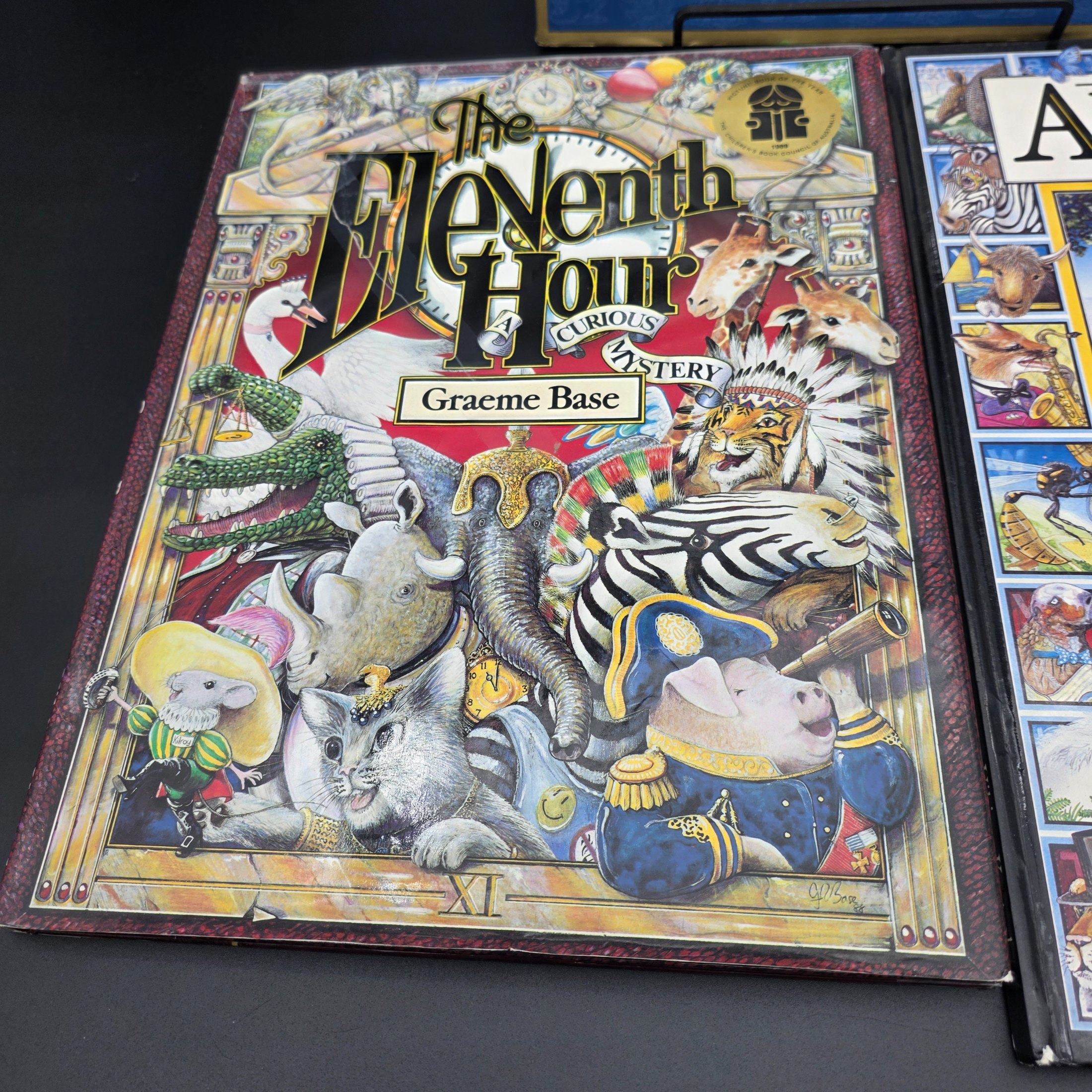 A collection of three hardcover children's books by Graeme Base: 'The Discovery of Dragons', 'The Eleventh Hour Curious Mystery', and 'Animalia', featuring intricate illustrations. - View 3