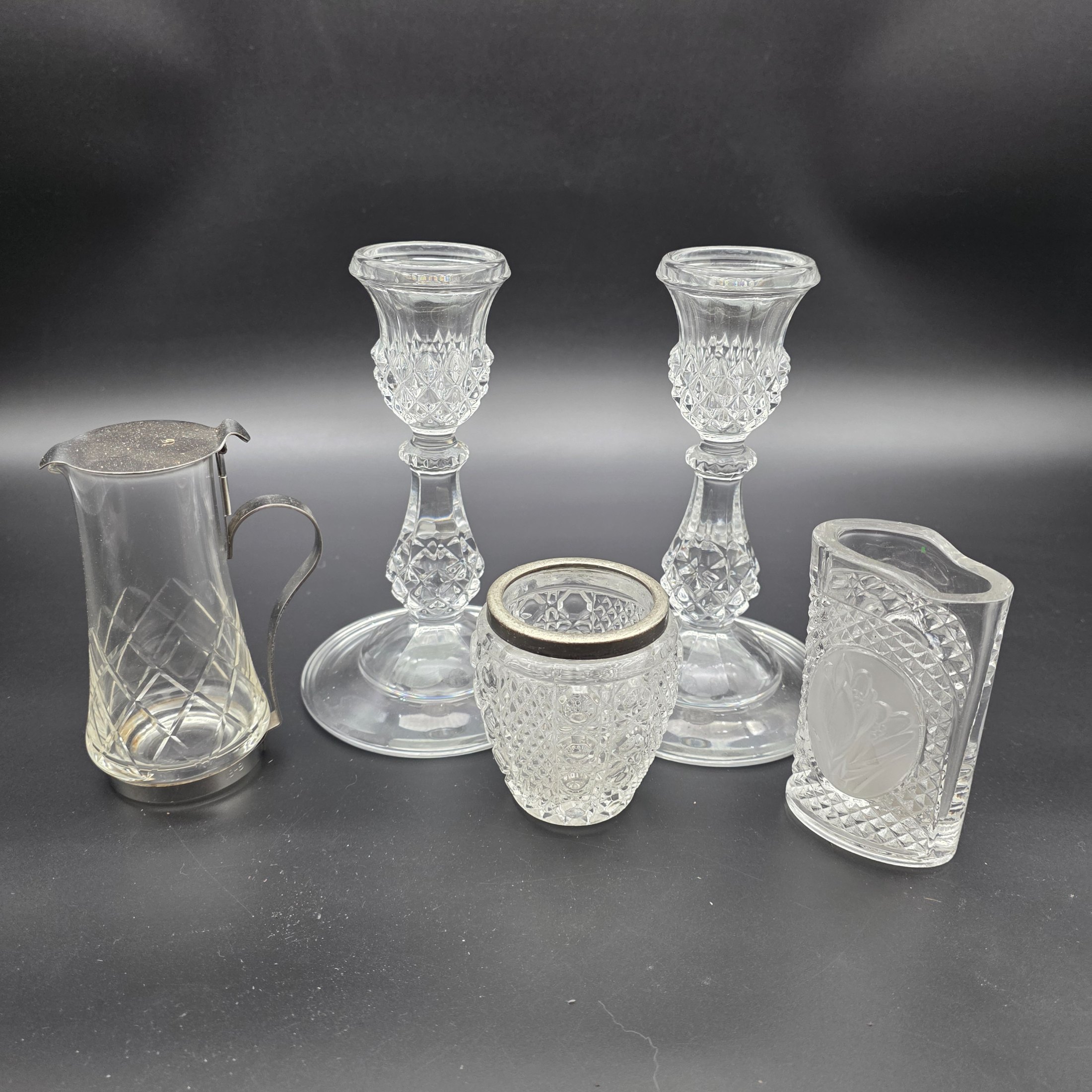 A diverse collection of clear and frosted pressed glass decorative items, comprising a pair of candlesticks, a small pitcher with metal handle and lid, a textured jar with a metal rim, and a rectangular vase with a frosted floral motif, likely from the early to mid-20th century.