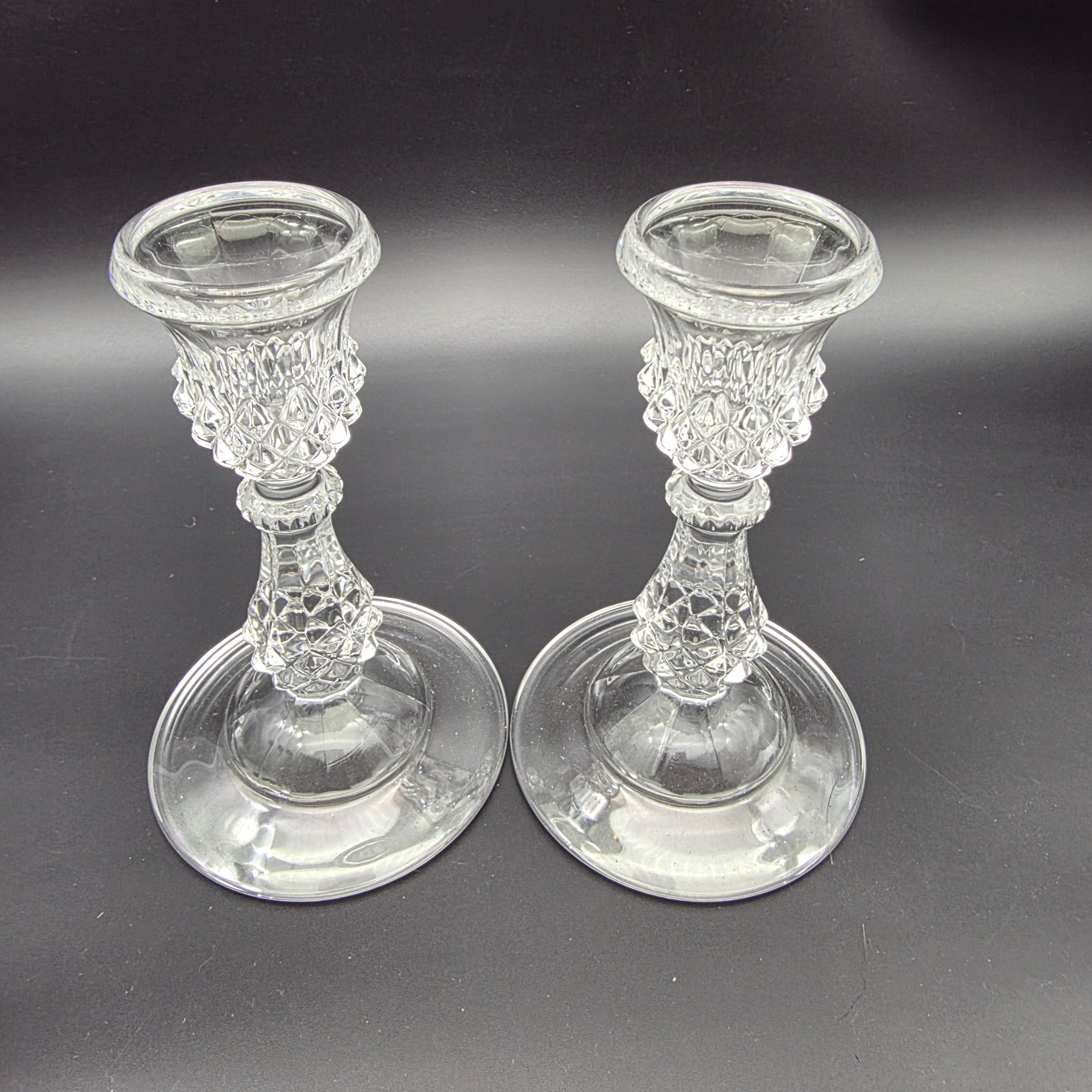 A diverse collection of clear and frosted pressed glass decorative items, comprising a pair of candlesticks, a small pitcher with metal handle and lid, a textured jar with a metal rim, and a rectangular vase with a frosted floral motif, likely from the early to mid-20th century. - View 3