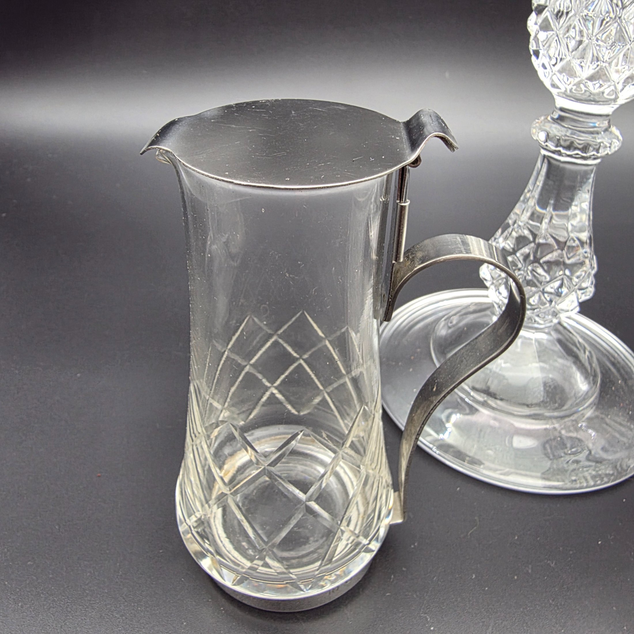 A diverse collection of clear and frosted pressed glass decorative items, comprising a pair of candlesticks, a small pitcher with metal handle and lid, a textured jar with a metal rim, and a rectangular vase with a frosted floral motif, likely from the early to mid-20th century. - View 5