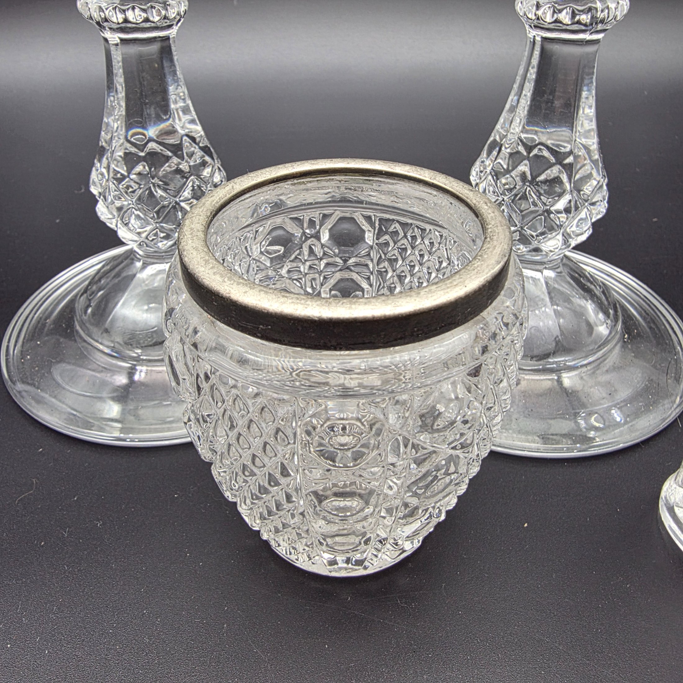 A diverse collection of clear and frosted pressed glass decorative items, comprising a pair of candlesticks, a small pitcher with metal handle and lid, a textured jar with a metal rim, and a rectangular vase with a frosted floral motif, likely from the early to mid-20th century. - View 7