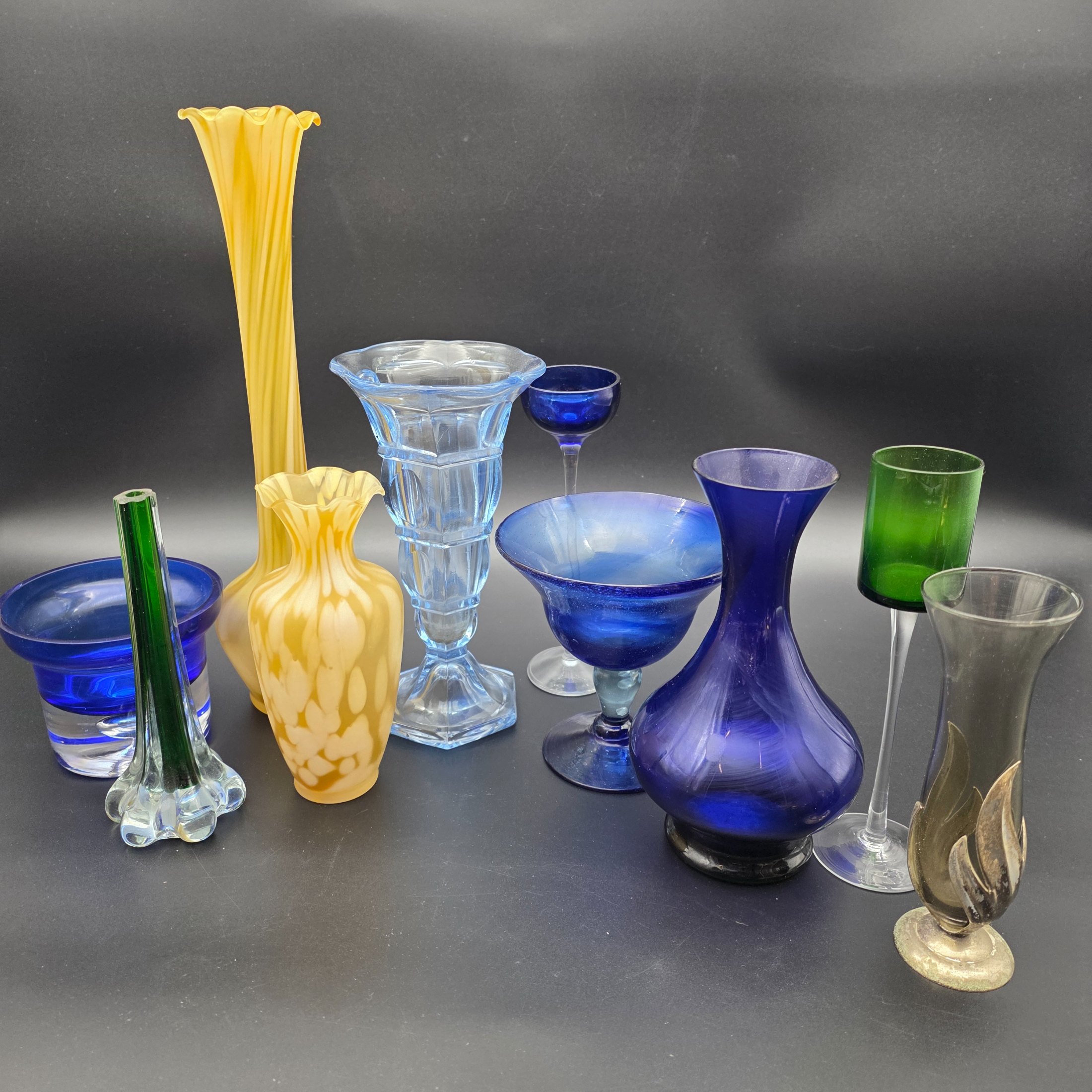 A varied collection of ten vintage coloured glass pieces, including vases, bowls, and stemmed glasses, showcasing different styles and manufacturing techniques from the mid-20th century. The array features shades of cobalt blue, amber, yellow, green, and smoky grey, offering a diverse decorative grouping.