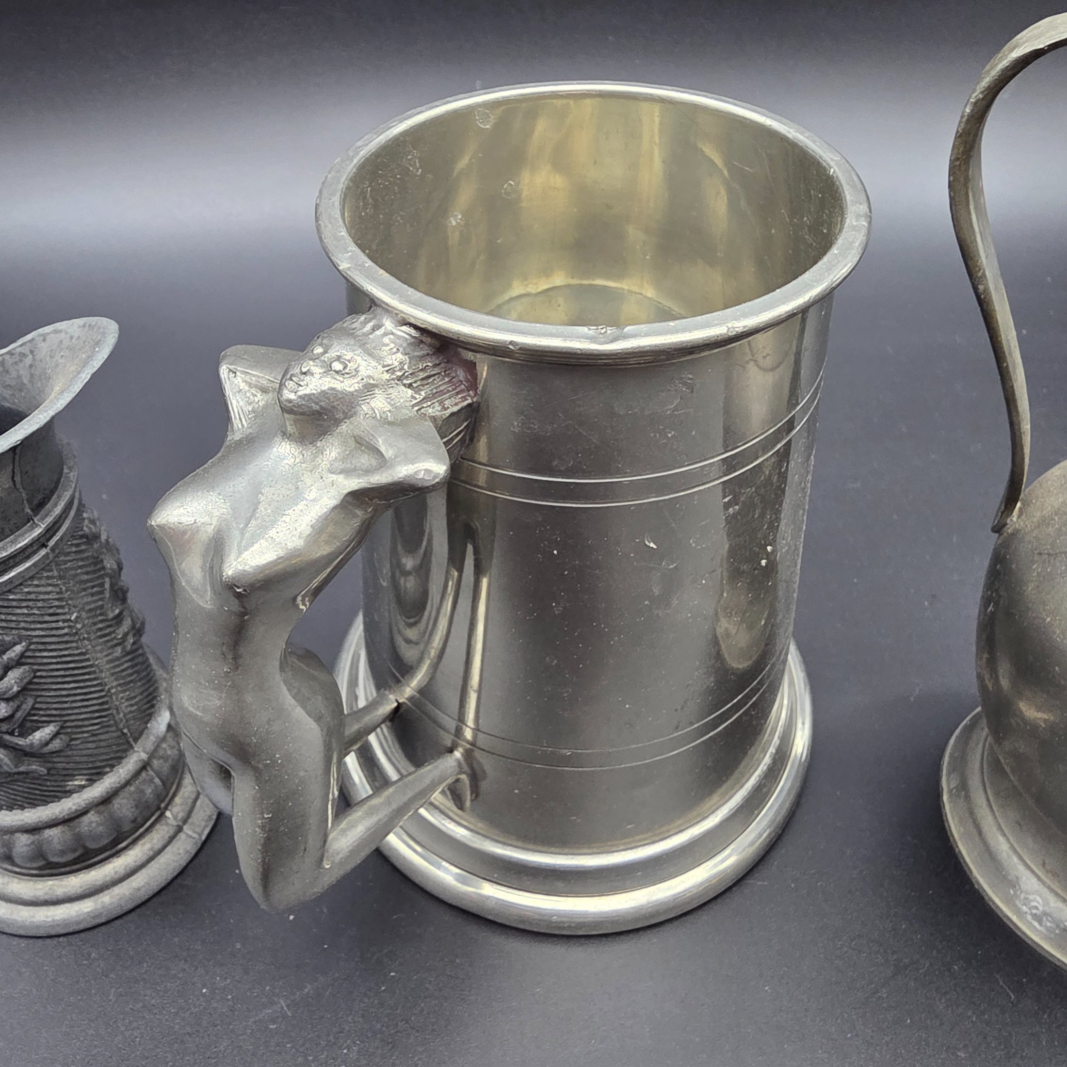 A collection of three traditional English pewter vessels, including a tankard with a unique nude figure handle, and two pitchers. These pieces originate from Sheffield, a city historically renowned for its metalwork and pewter manufacturing.