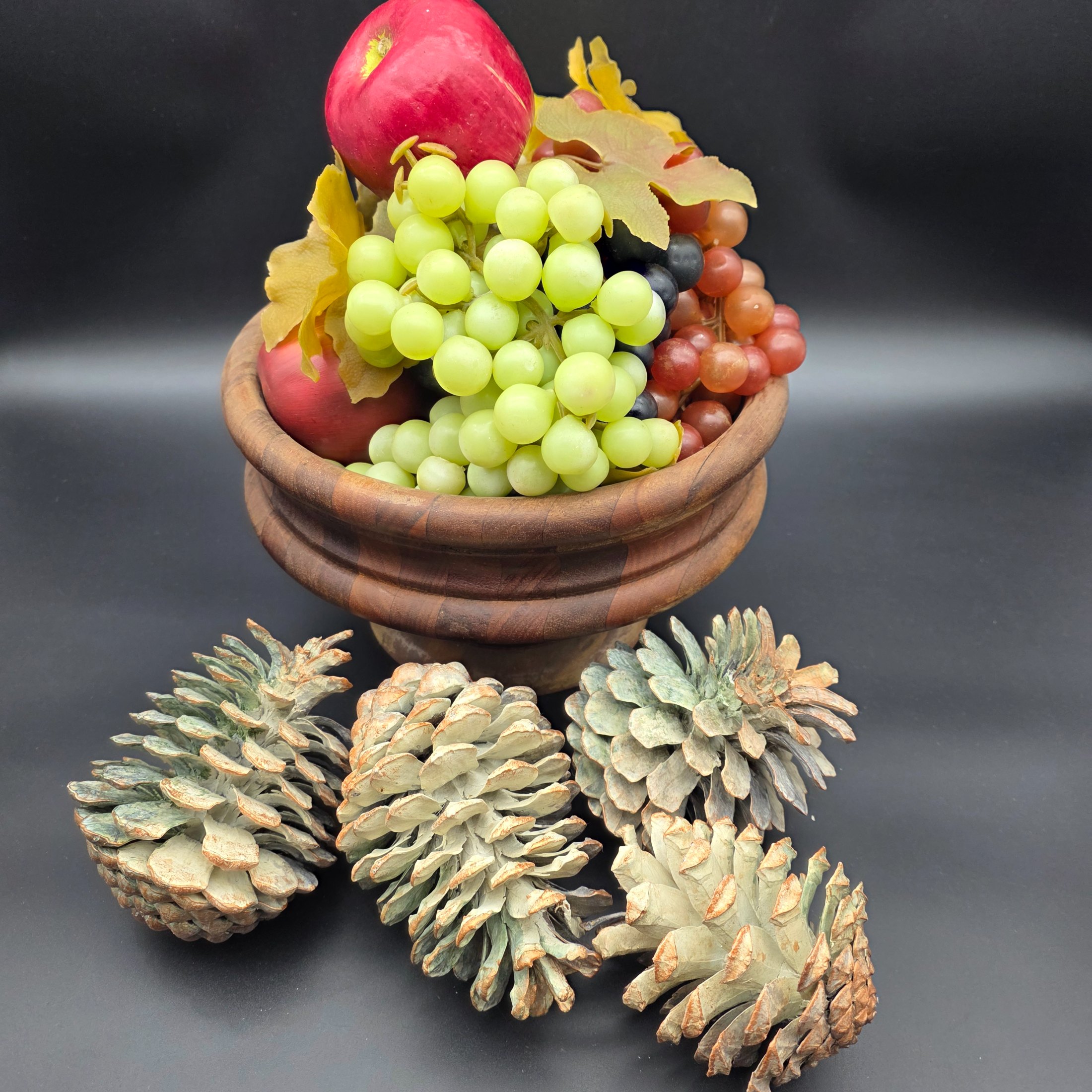 A decorative ensemble featuring a turned wooden bowl filled with artificial fruit (apples and grapes) and faux autumn leaves, accompanied by several large natural pinecones.
