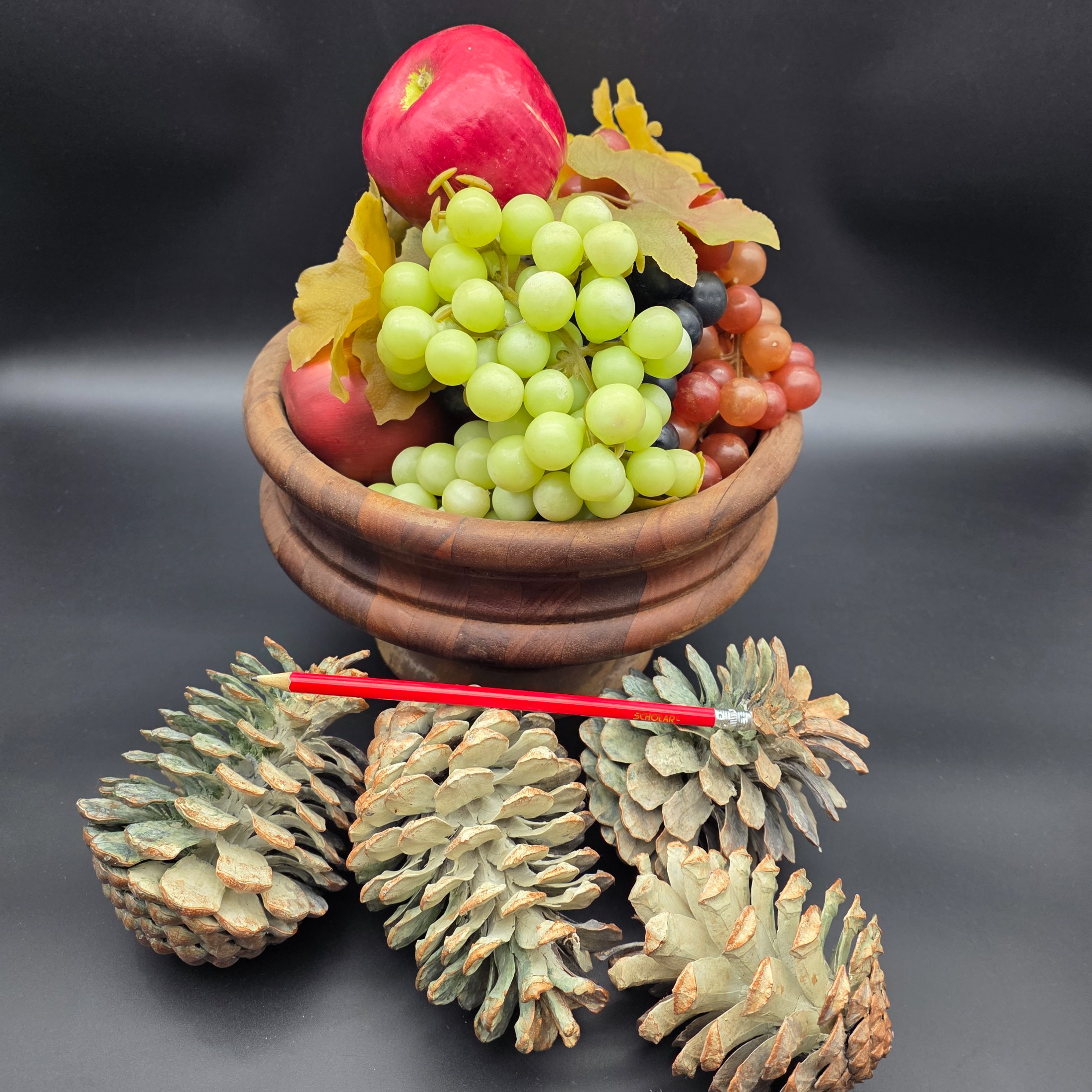 A decorative ensemble featuring a turned wooden bowl filled with artificial fruit (apples and grapes) and faux autumn leaves, accompanied by several large natural pinecones. - View 2